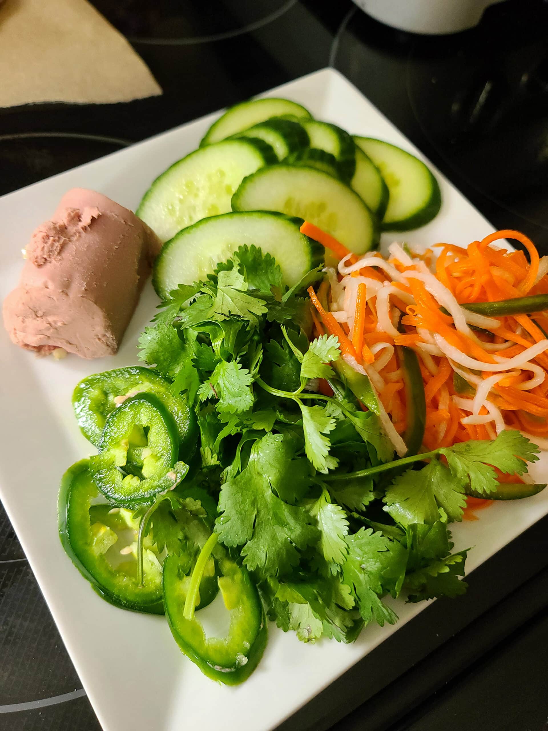 A plate with cucumber slices, picked carrots and daikon radish, cilantro, jalapeno slices, and liver pate.