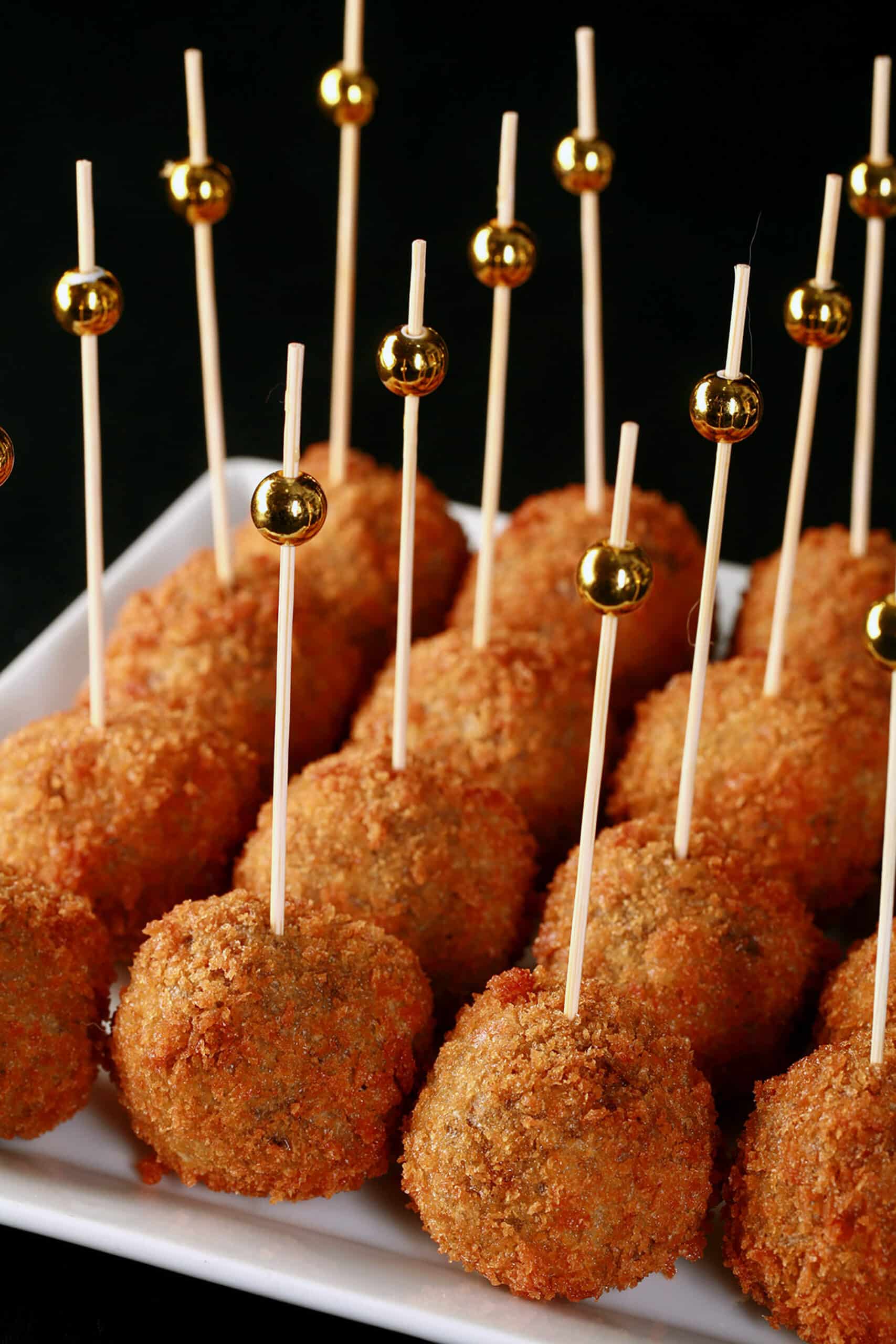 16 mini mushroom arancini on a plate, each with a tall toothpick topped with a gold ball.