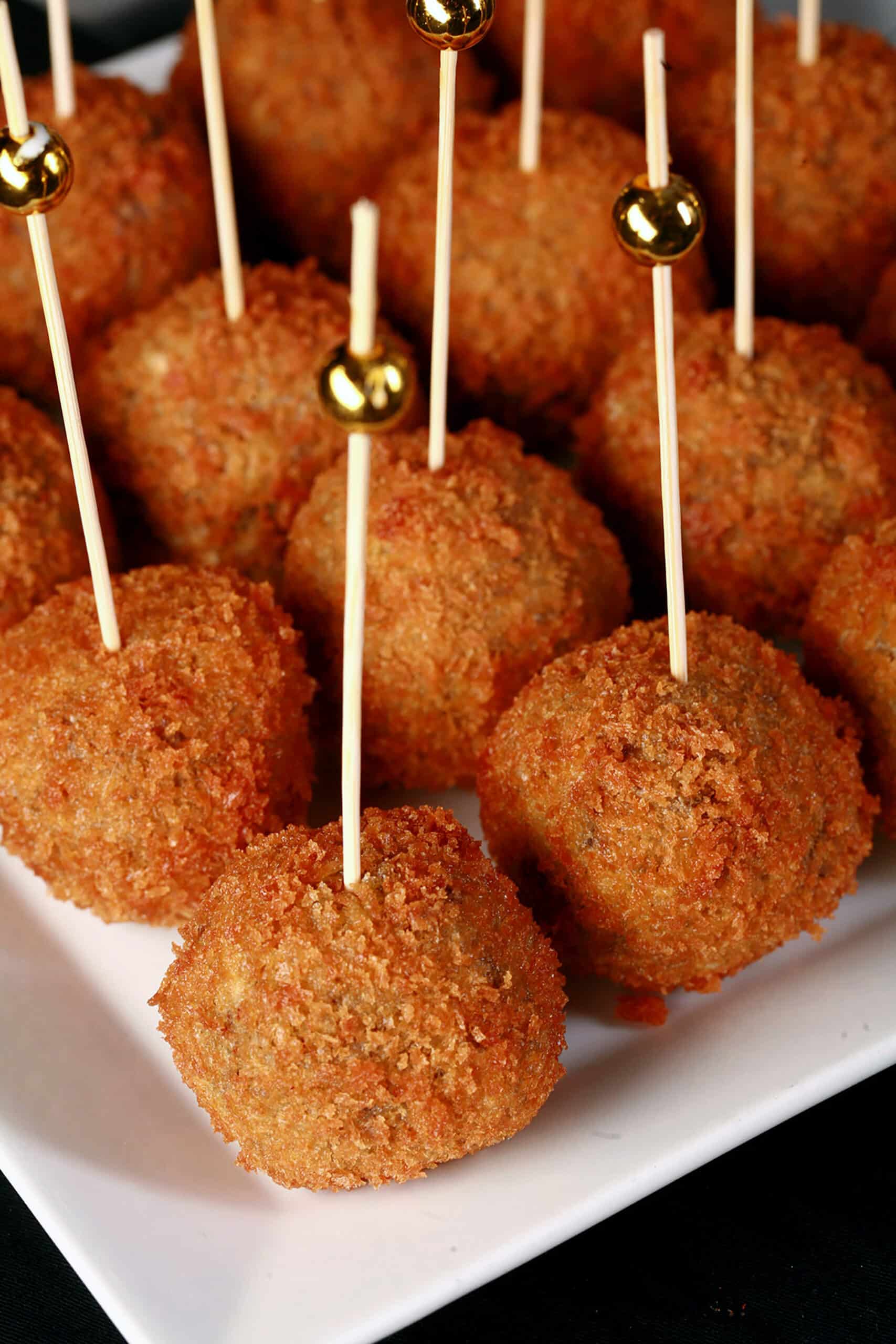 16 mini mushroom risotto balls on a plate, each with a tall toothpick topped with a gold ball.
