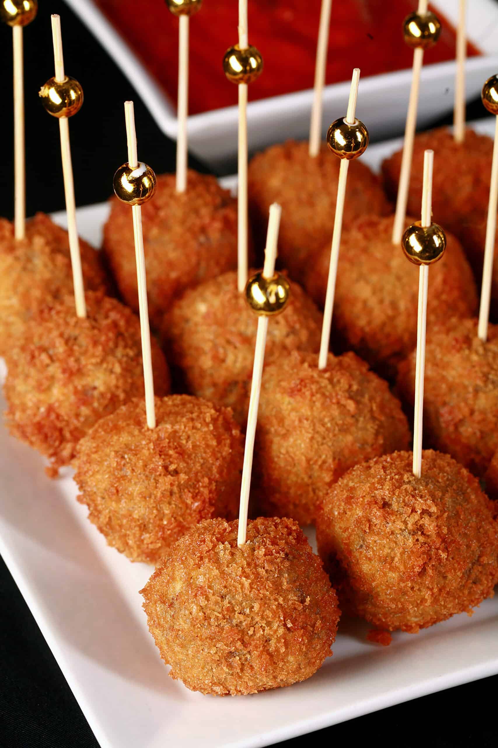 16 mini mushroom risotto balls on a plate, each with a tall toothpick topped with a gold ball.   There is a small bowl of marinara behind the plate.