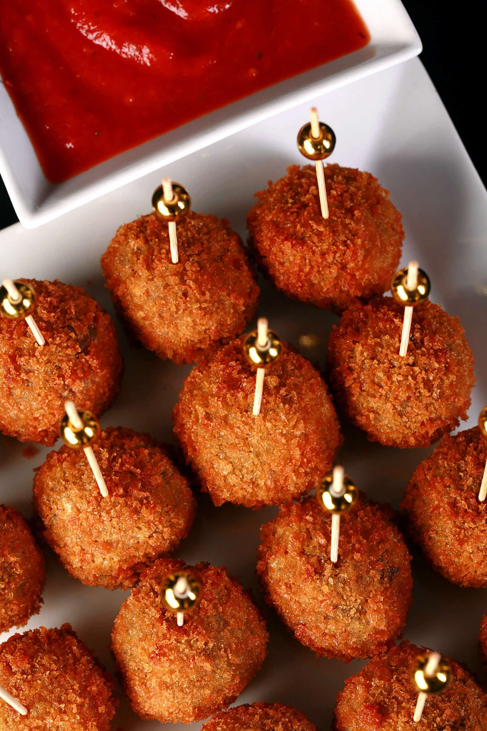 16 mini mushroom arancini on a plate, each with a tall toothpick topped with a gold ball. There is a small bowl of marinara behind the plate.