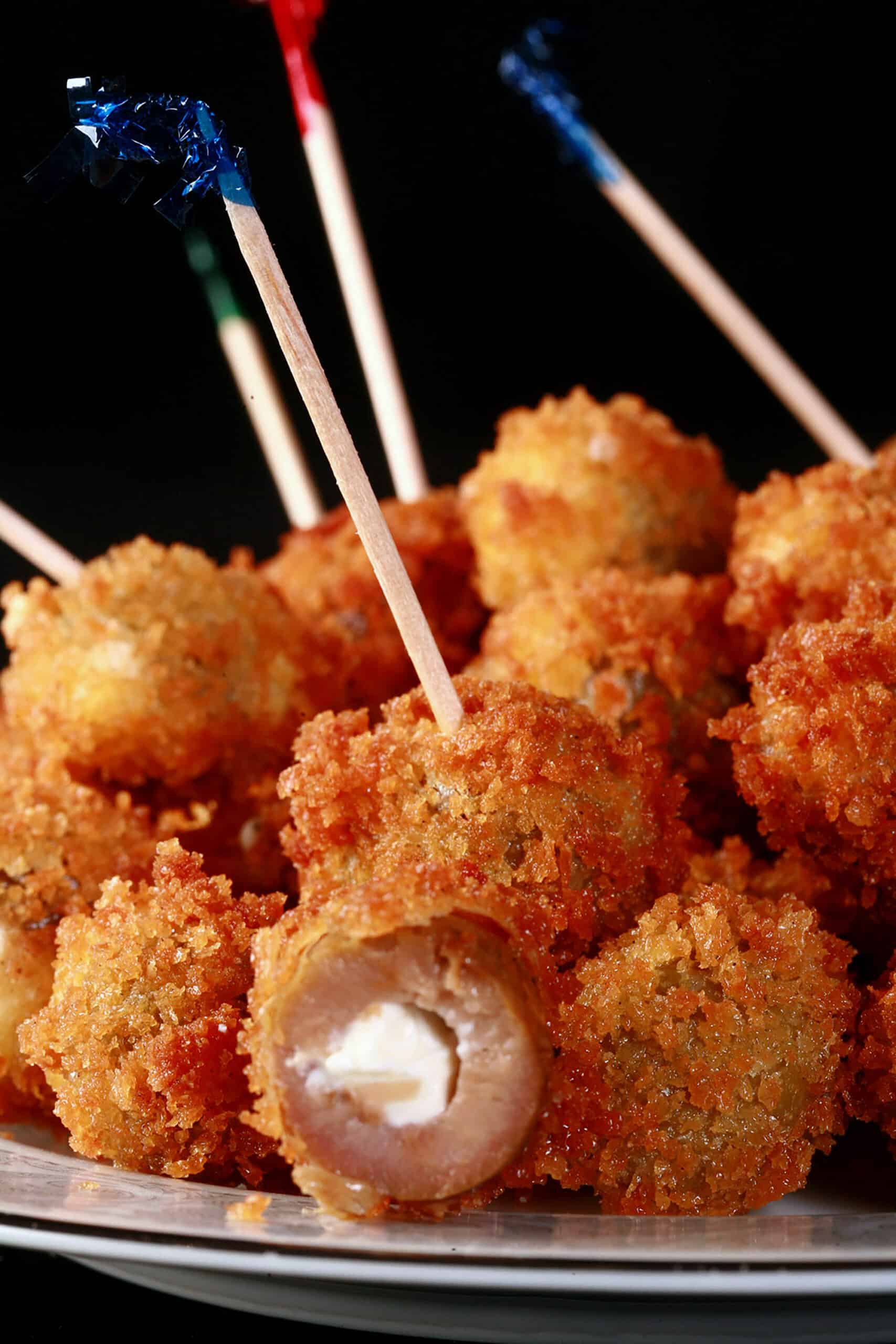 A pile of breaded deep fried olives on a plate. One is broken open to show it’s stuffed with a garlic cheese mixture.