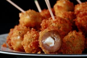 A pile of breaded deep fried olives on a plate. One is broken open to show it's stuffed with a garlic cheese mixture.
