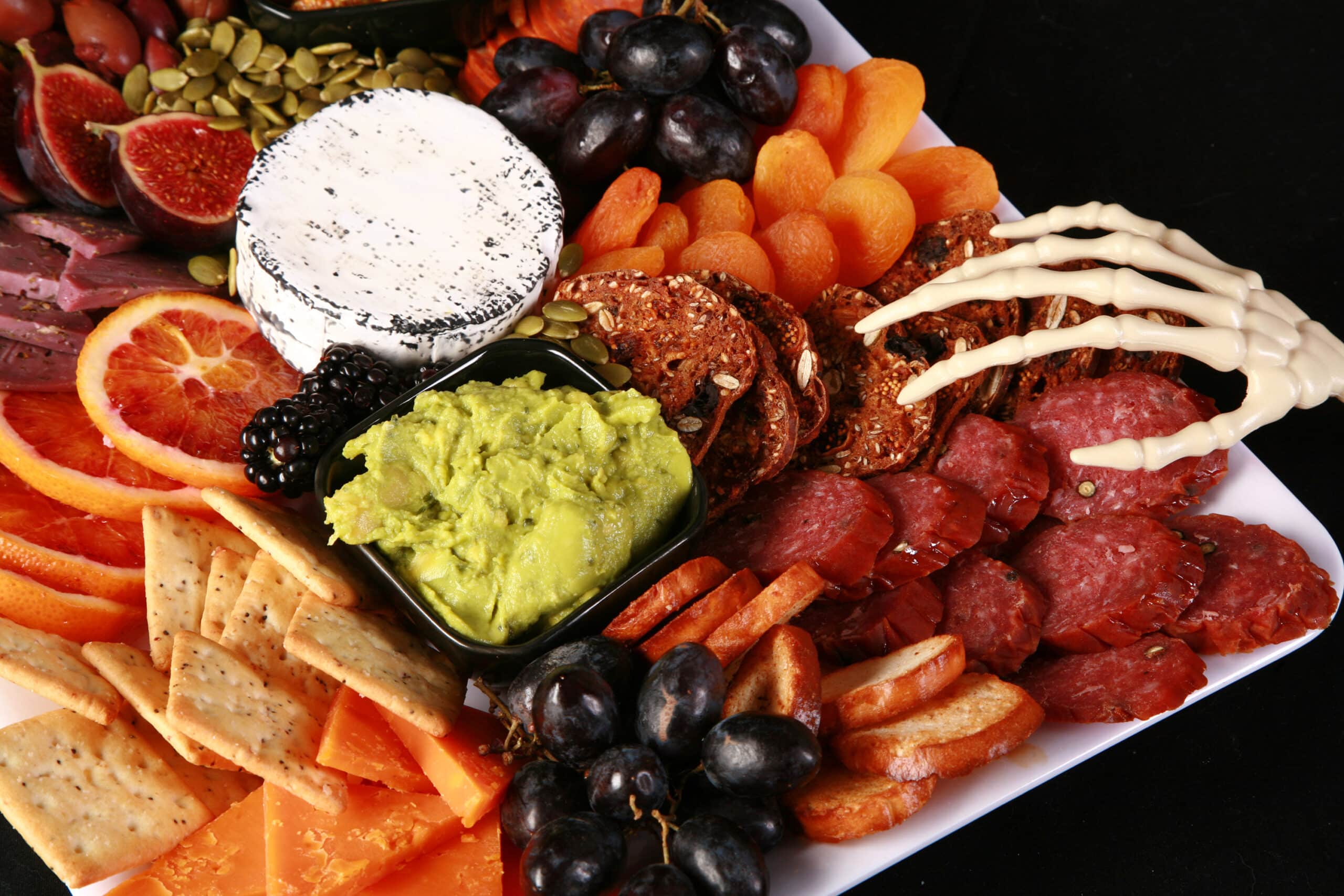 An spooky halloween charcuterie board with jewel toned foods and skeleton hand accents.