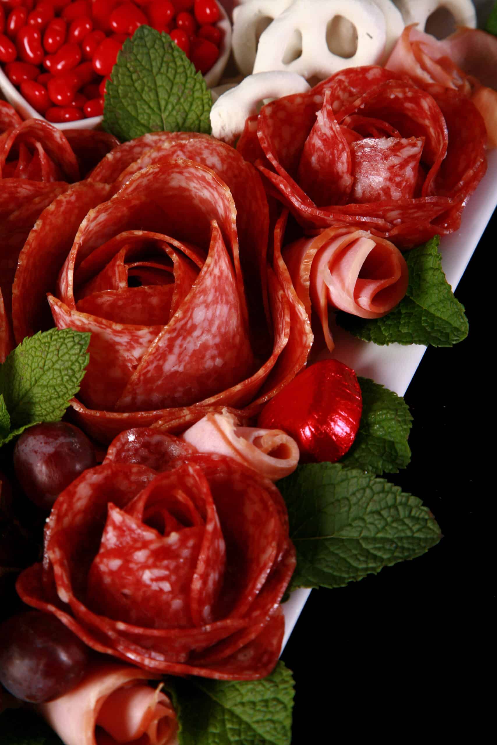 A valentine’s day charcuterie board, with heart shaped cheese slices, salami roses, foil wrapped chocolates, strawberries, and more.