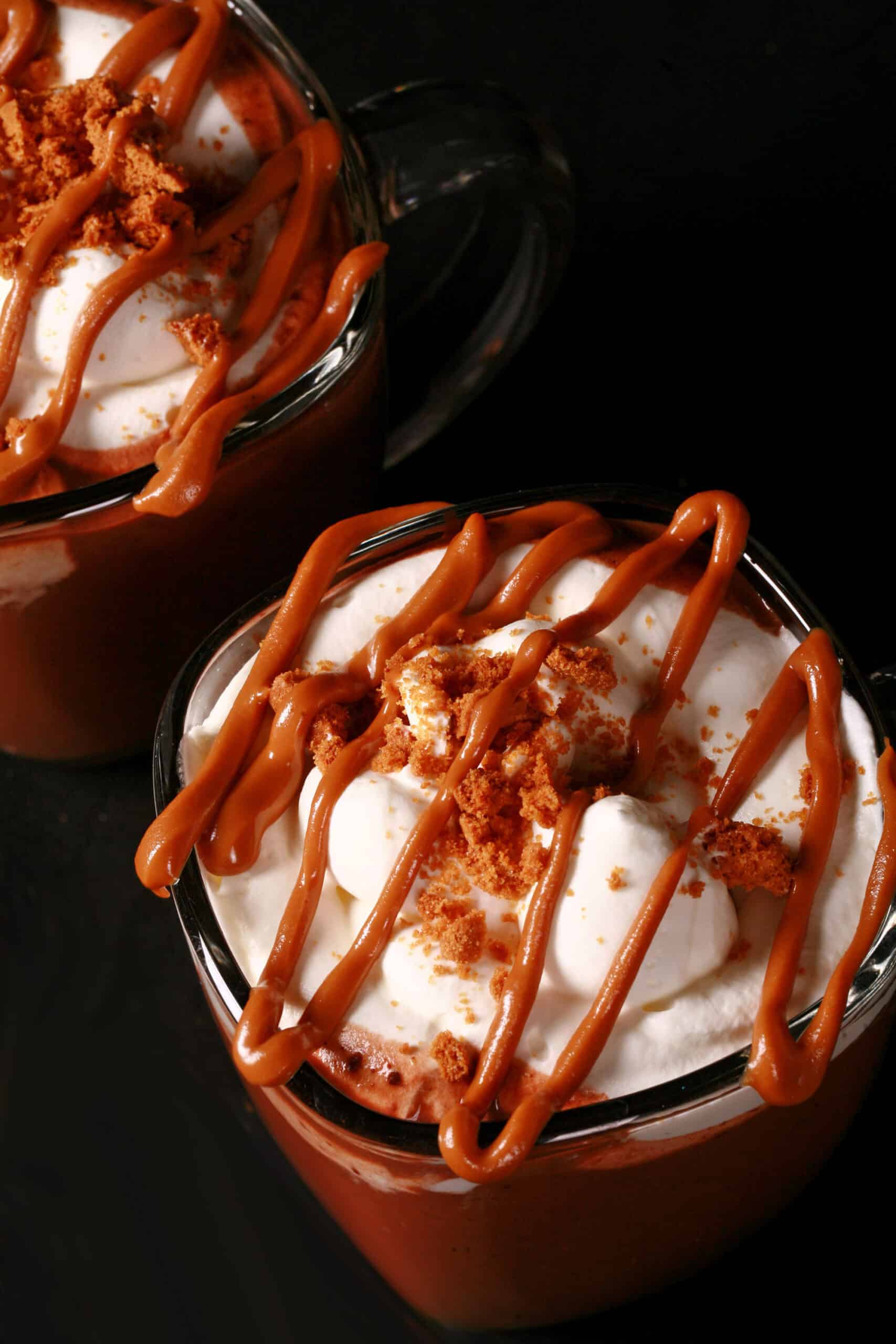 2 mugs of Biscoff hot chocolate, garnished with whipped cream, drizzled cookie butter, and crushed biscoff cookies.