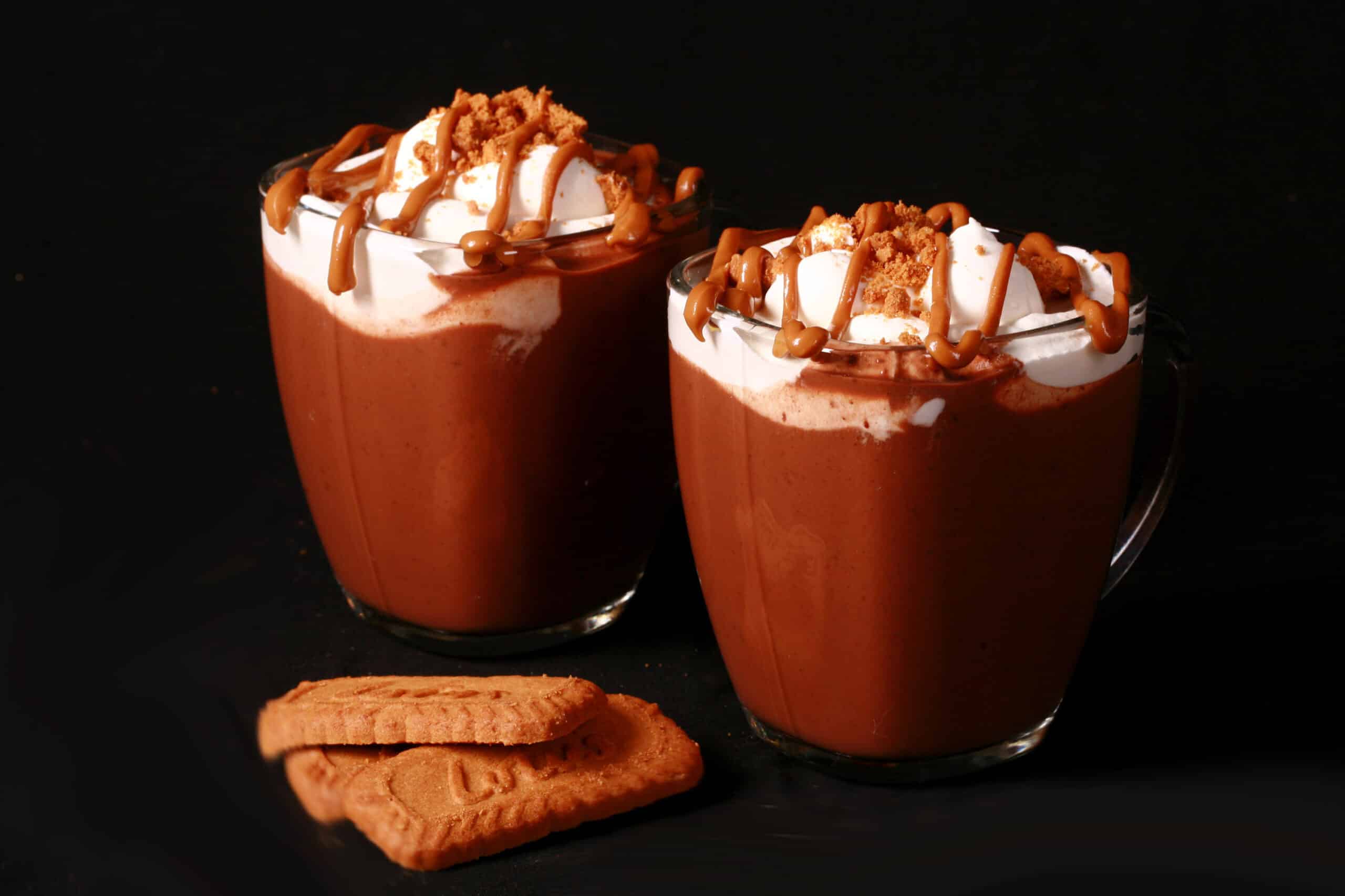 2 mugs of Biscoff hot cocoa, garnished with whipped cream, drizzled cookie butter, and crushed biscoff cookies.