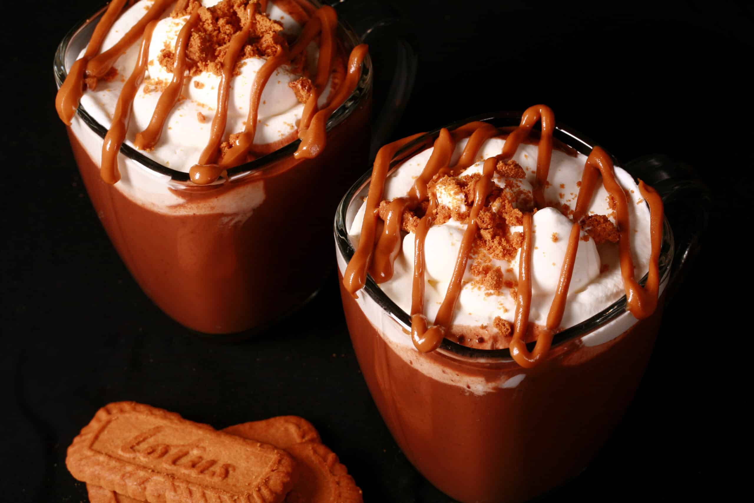 2 mugs of Biscoff hot chocolate, garnished with whipped cream, drizzled cookie butter, and crushed biscoff cookies.