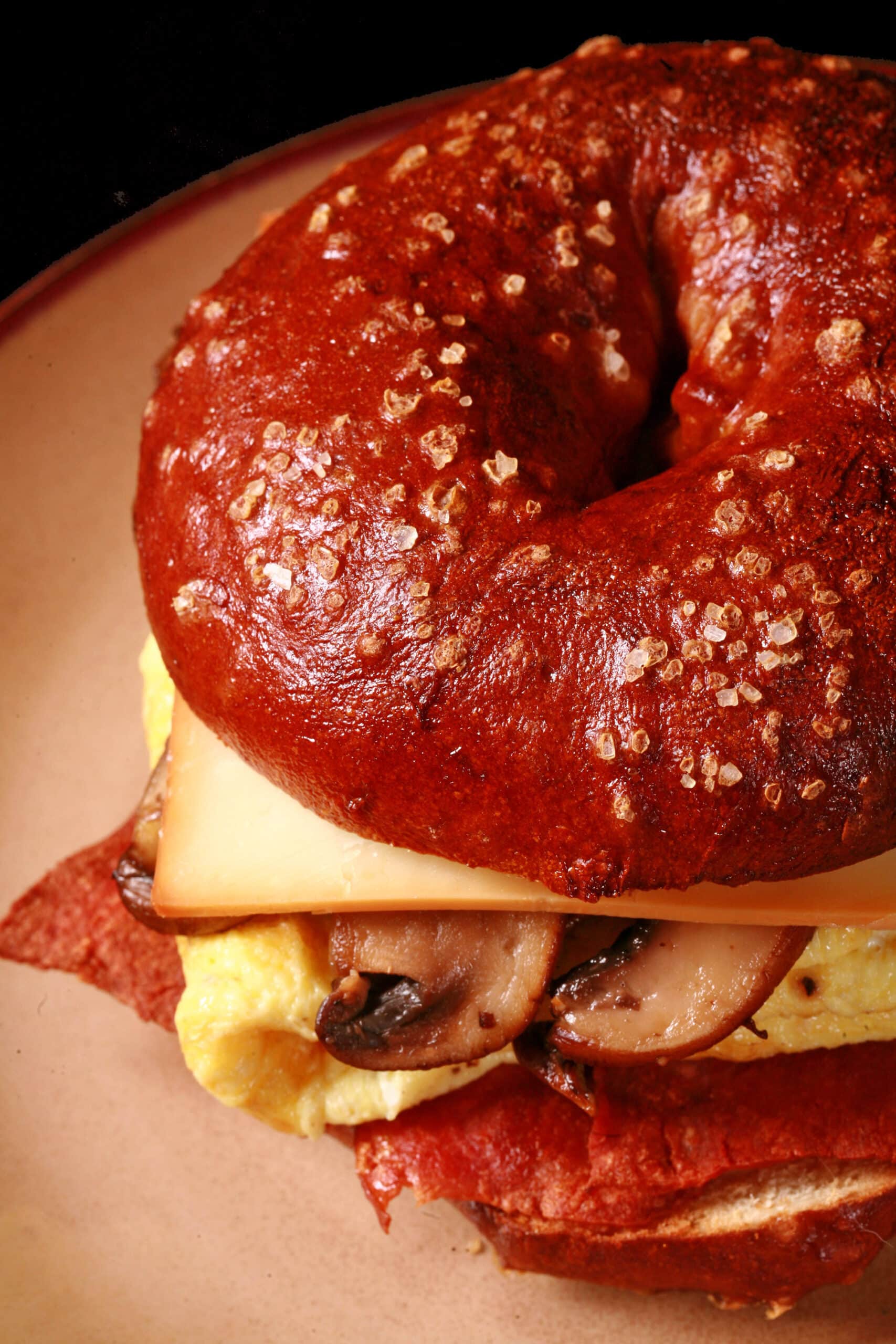 A breakfast sandwich made with a pretzel bagel, omelette, mushrooms, bacon, and cheese.