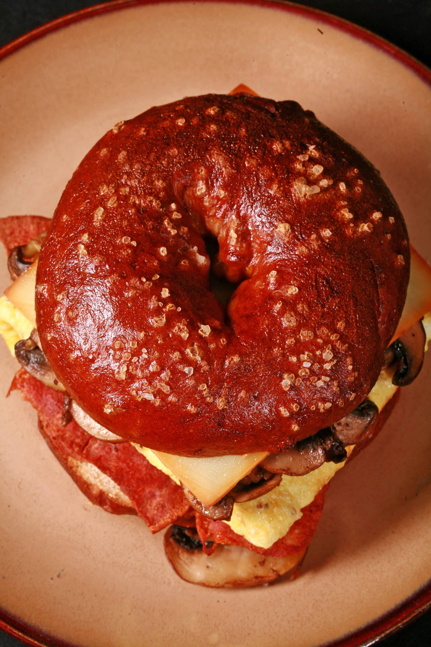 A breakfast sandwich made with a pretzel bagel, omelette, mushrooms, bacon, and cheese.