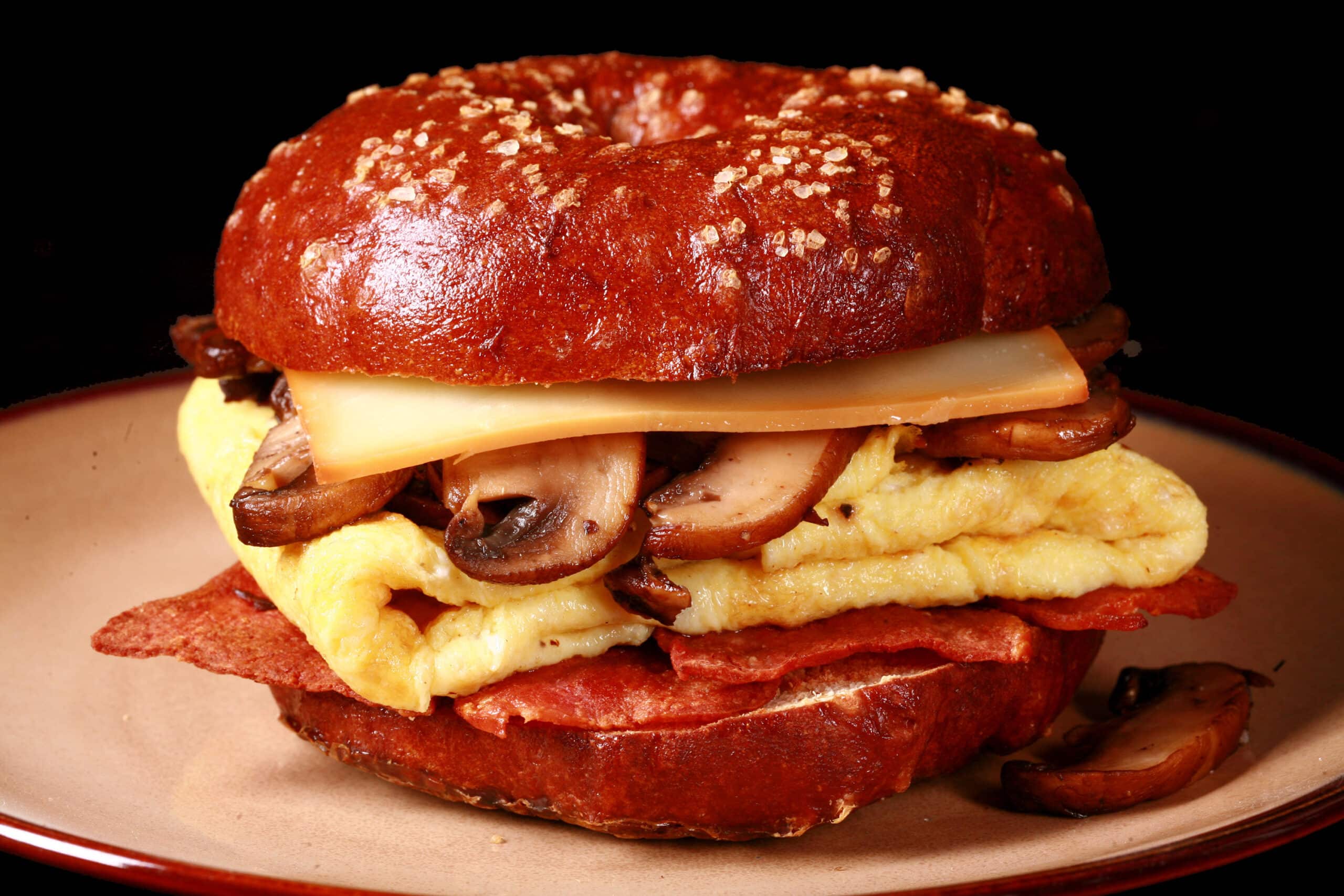 A breakfast sandwich made with a pretzel bagel, omelette, mushrooms, bacon, and cheese.