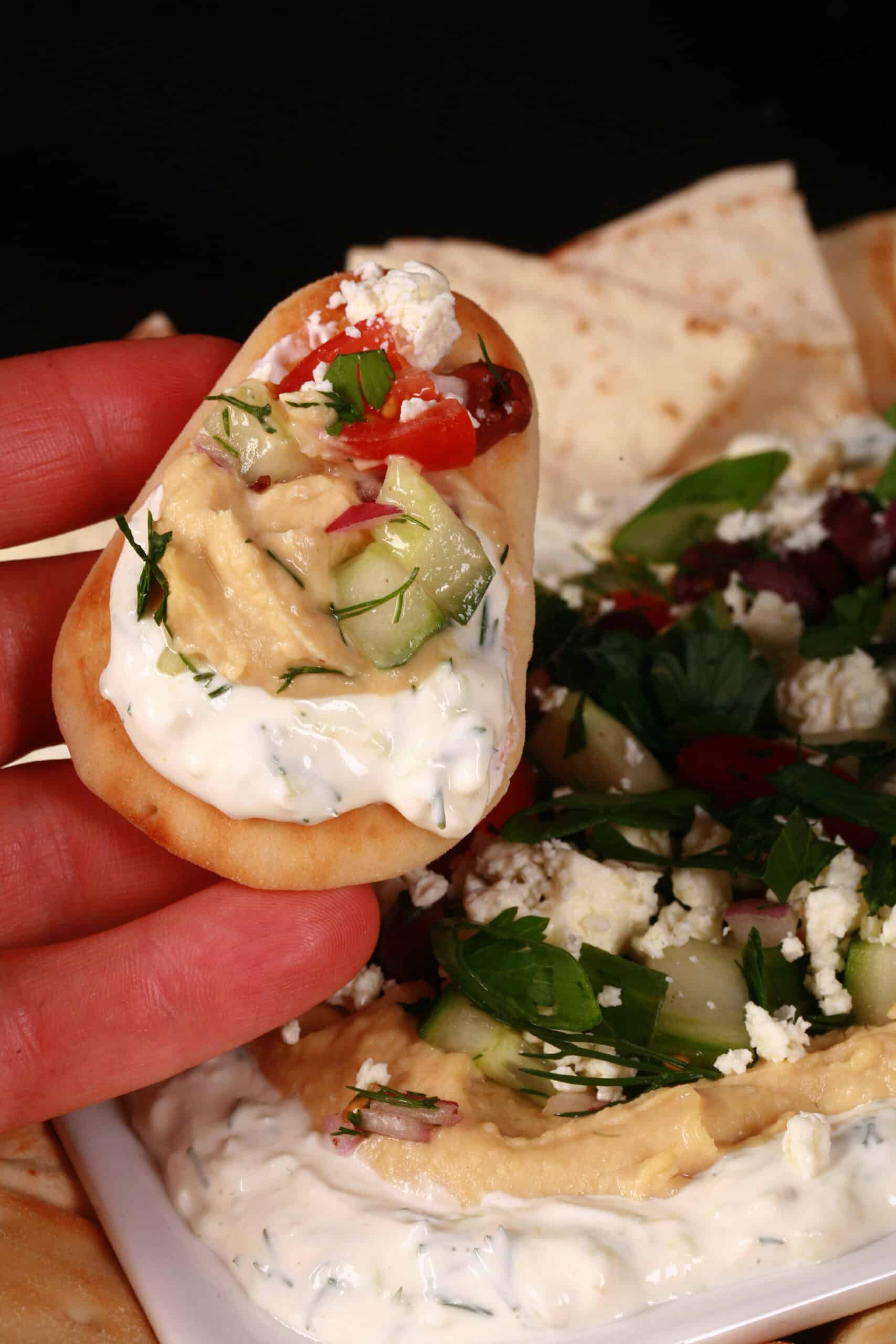 A hand holding a small dipping wedge of pita, with layered Greek dip on it.