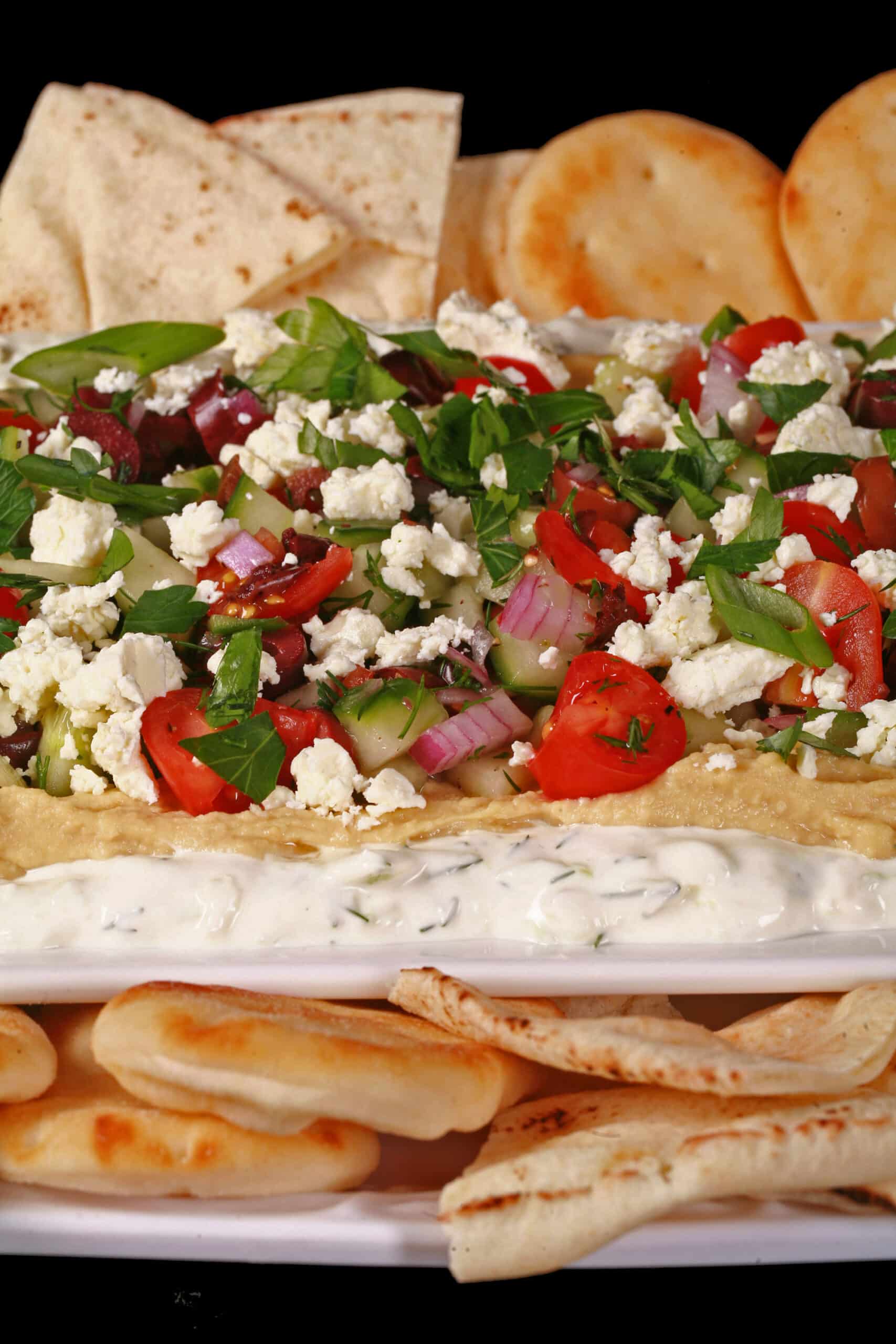 A plate of Greek Layered Dip, with Tzatziki cream cheese dip, hummus, a Greek salsa made of olives, cherry tomatoes, red onions, cucumber and fresh herbs, topped with feta.