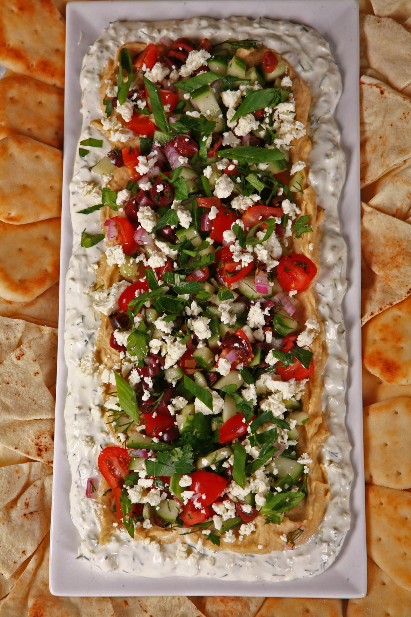 A plate of Greek Layered Dip, with Tzatziki cream cheese dip, hummus, a Greek salsa made of olives, cherry tomatoes, red onions, cucumber and fresh herbs, topped with feta.