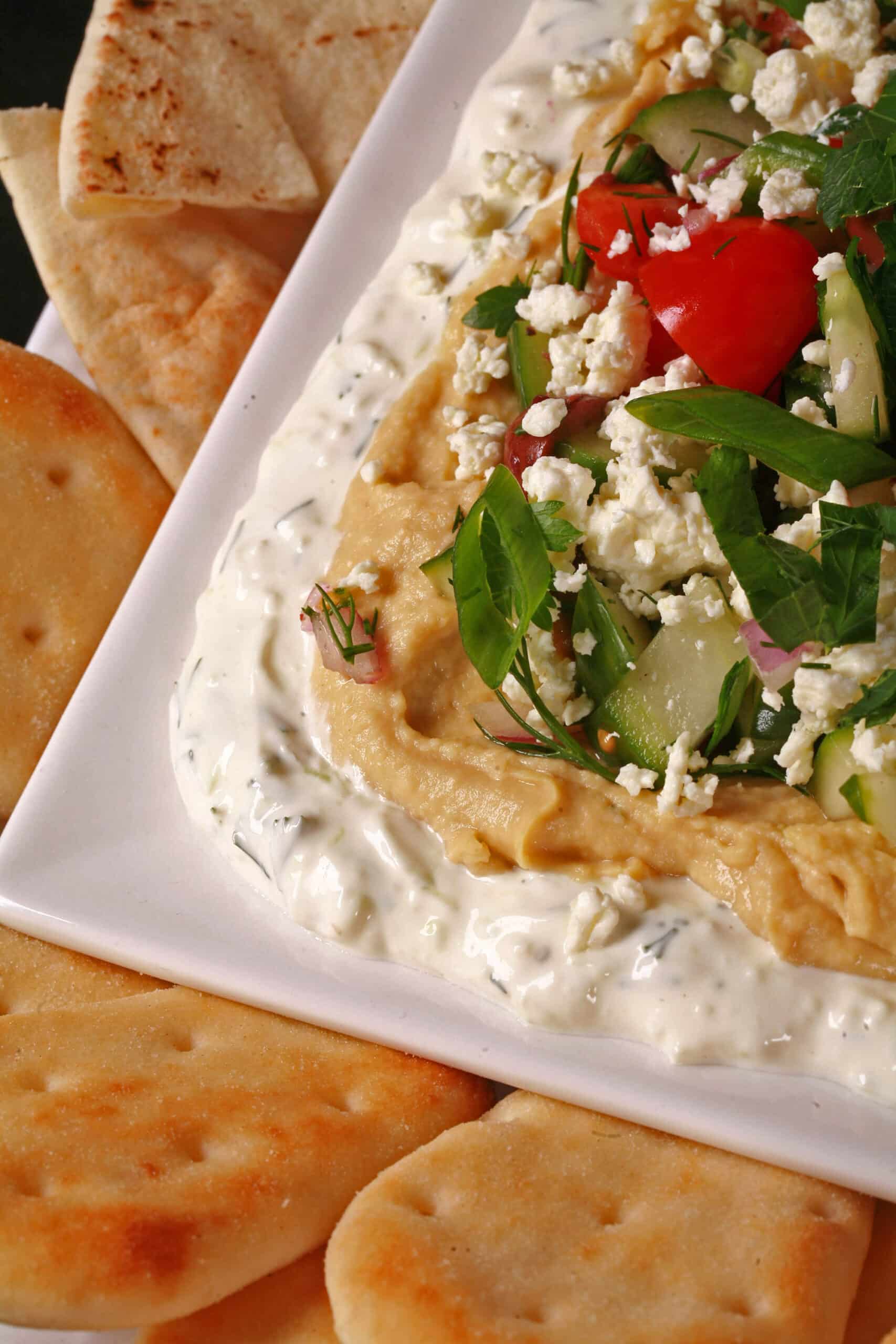 A plate of Layered Greek Dip, with Tzatziki cream cheese dip, hummus, a Greek salsa made of olives, cherry tomatoes, red onions, cucumber and fresh herbs, topped with feta.
