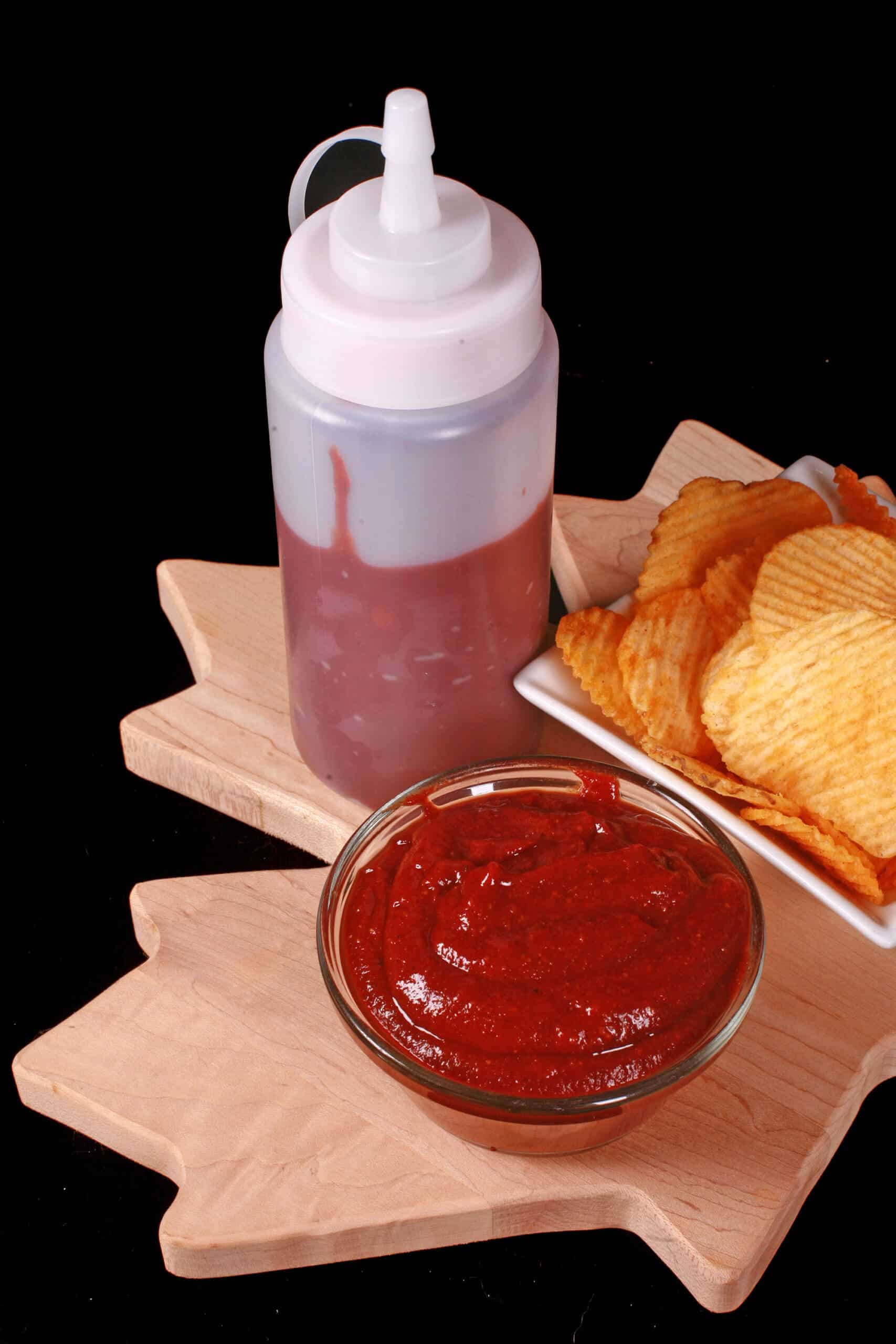 A small bowl of all dressed ketchup, in front of a bowl of all dressed chips and a bottle of ketchup.