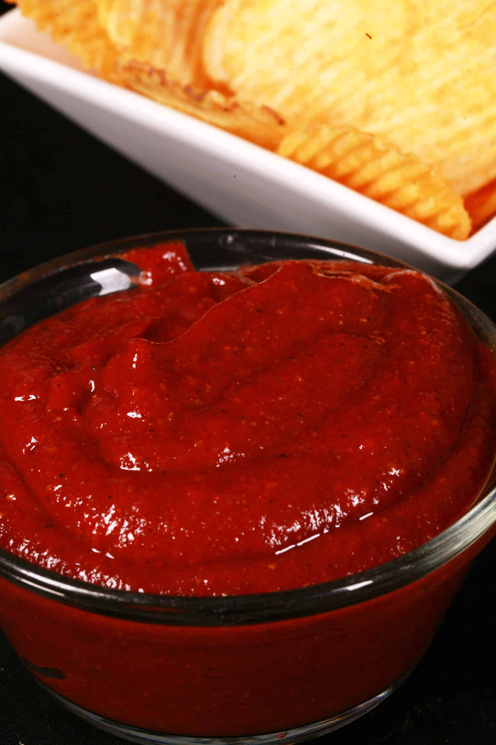 A small bowl of all dressed ketchup, in front of a bowl of all dressed chips.