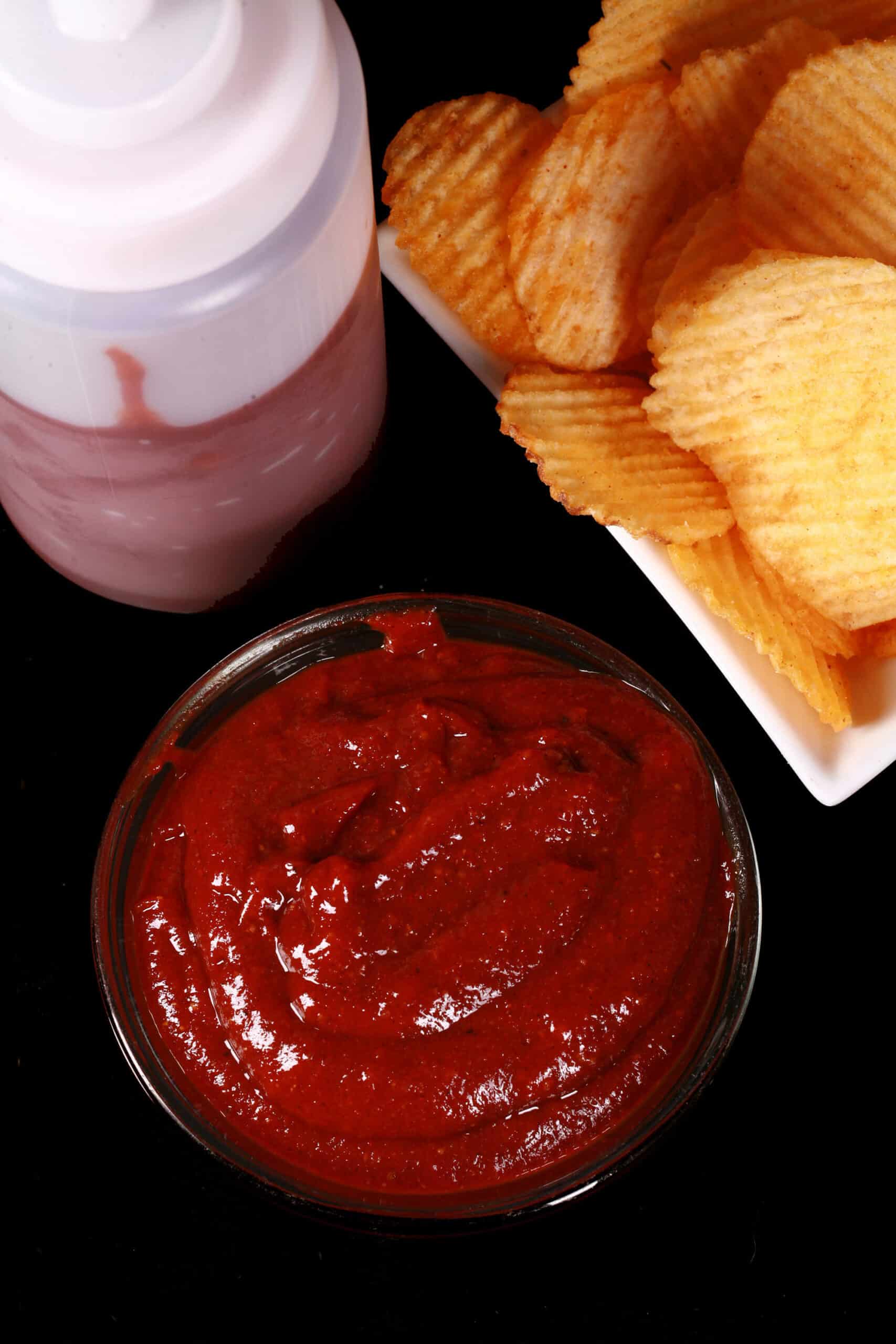A small bowl of all dressed ketchup, in front of a bowl of all dressed chips and a bottle of ketchup.