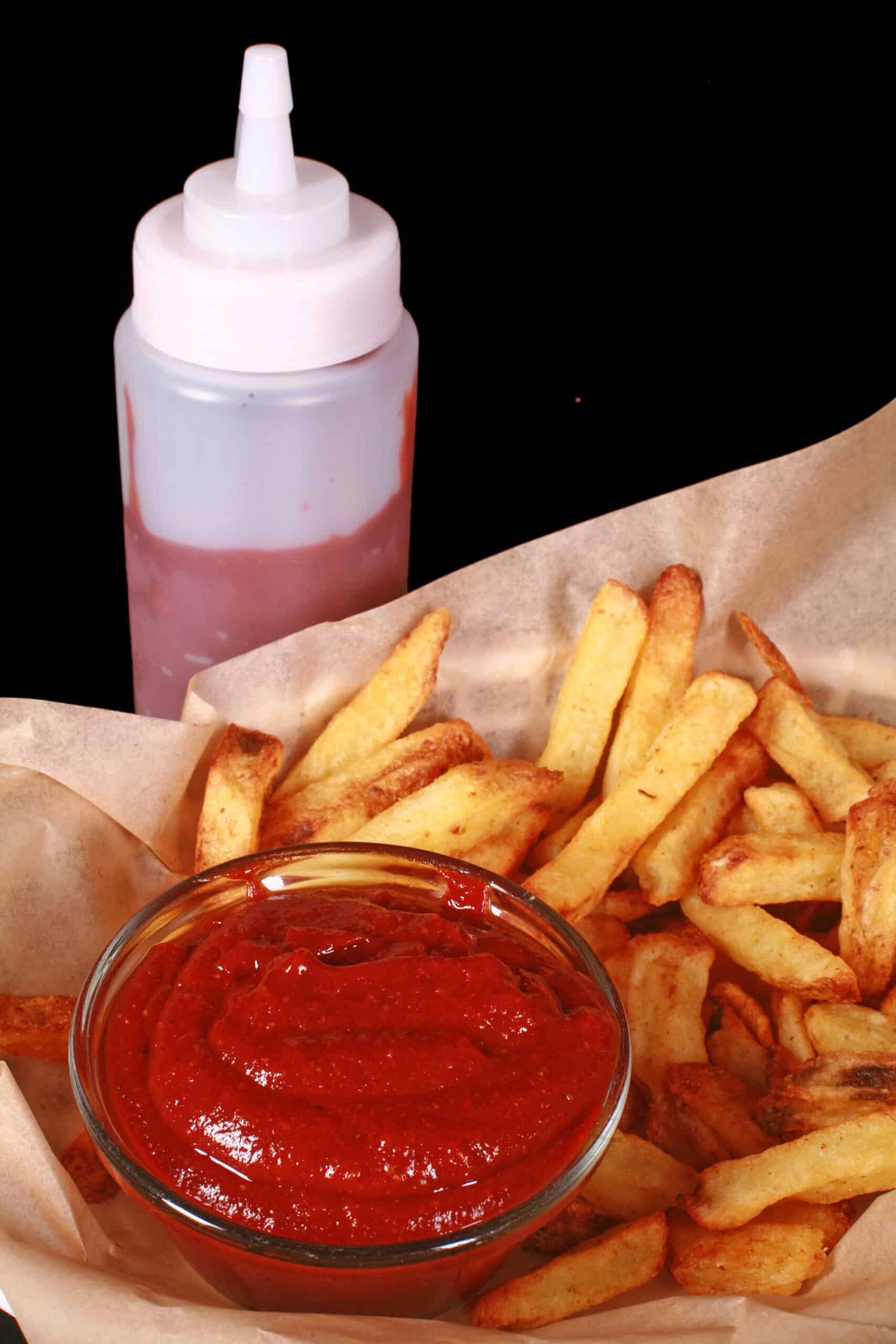 A small bowl of all dressed ketchup, in a bowl of fries, next to a bottle of ketchup.