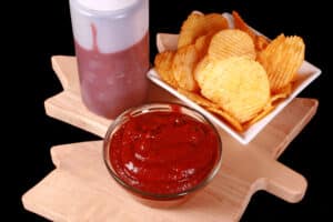 A small bowl of all dressed ketchup, in front of a bowl of all dressed chips and a bottle of ketchup.