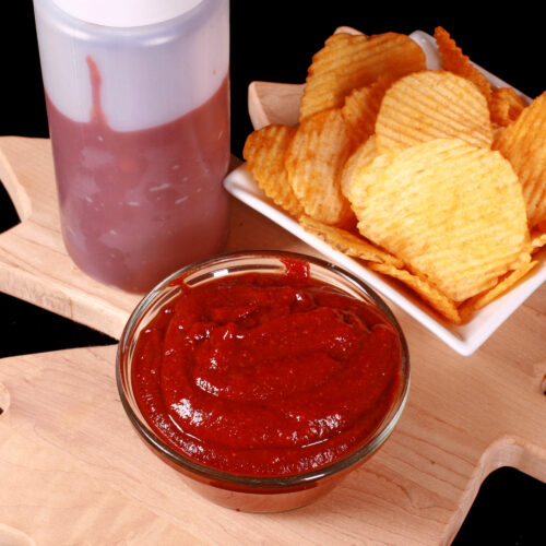 A small bowl of all dressed ketchup, in front of a bowl of all dressed chips and a bottle of ketchup.