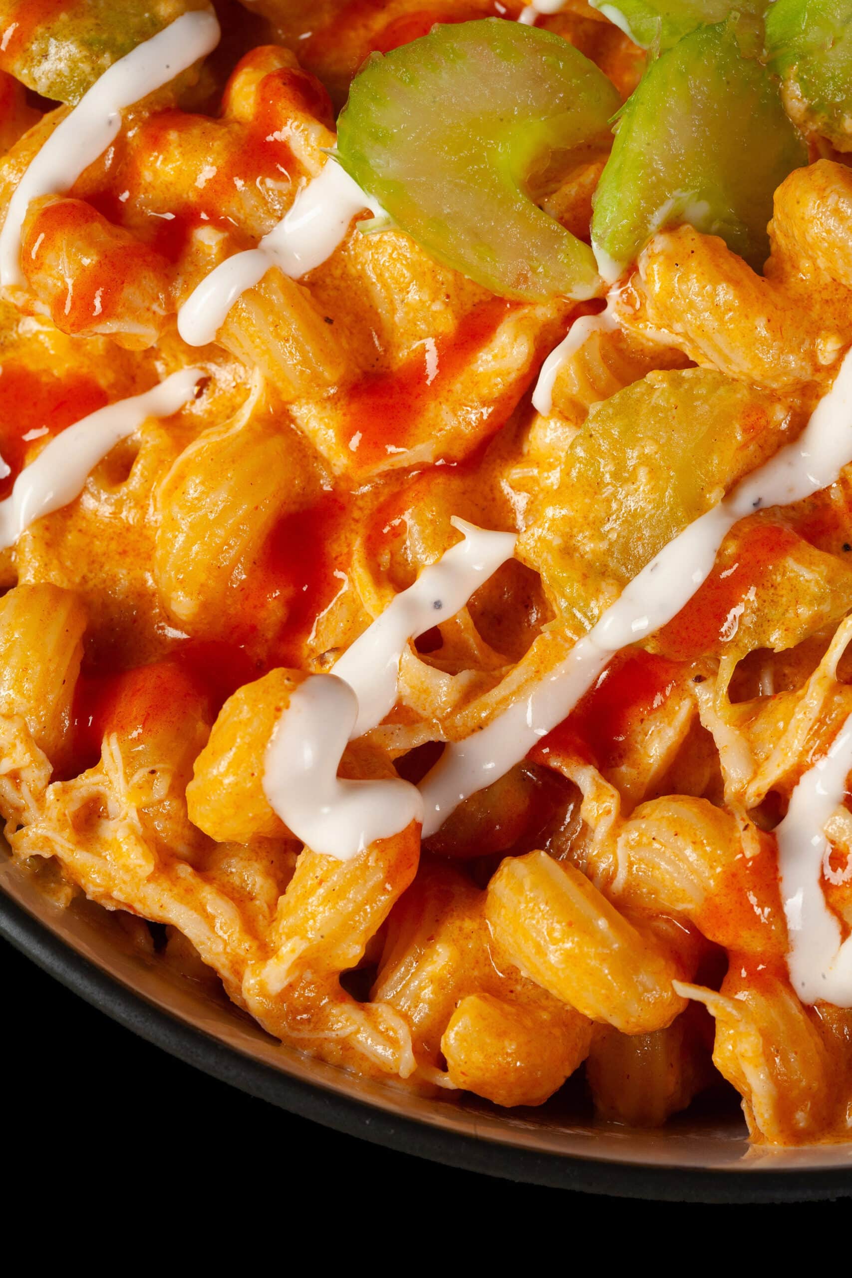 A bowl of buffalo chicken mac and cheese, with spiral pasta, cheese sauce, shredded chicken, celery, and drizzles of ranch dressing and Frank’s redhot sauce.