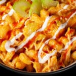 A bowl of buffalo chicken mac and cheese, with spiral pasta, cheese sauce, shredded chicken, celery, and drizzles of ranch dressing and frank's redhot sauce.