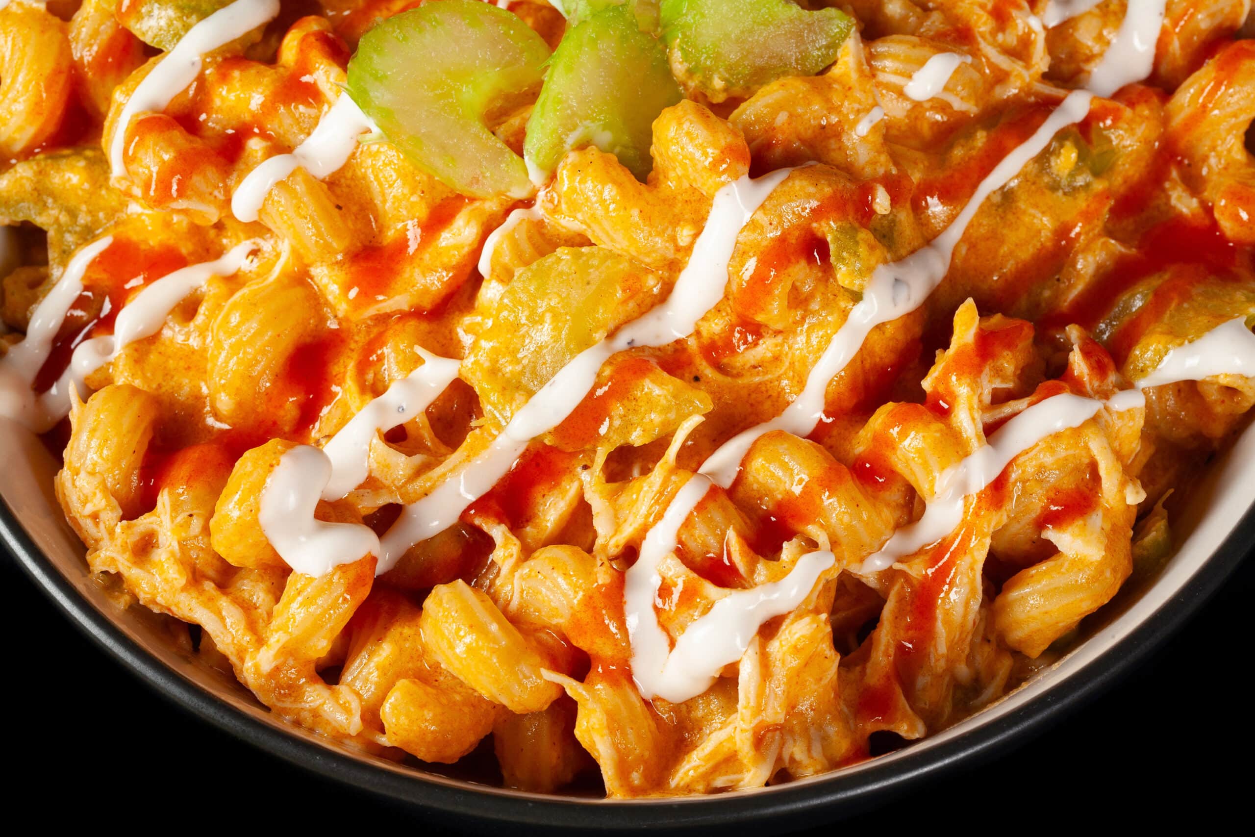 A bowl of buffalo chicken mac and cheese, with spiral pasta, cheese sauce, shredded chicken, celery, and drizzles of ranch dressing and Frank’s redhot sauce.