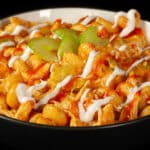 A bowl of buffalo chicken mac and cheese, with spiral pasta, cheese sauce, shredded chicken, celery, and drizzles of ranch dressing and frank's redhot sauce.