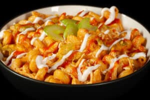 A bowl of buffalo chicken mac and cheese, with spiral pasta, cheese sauce, shredded chicken, celery, and drizzles of ranch dressing and frank's redhot sauce.