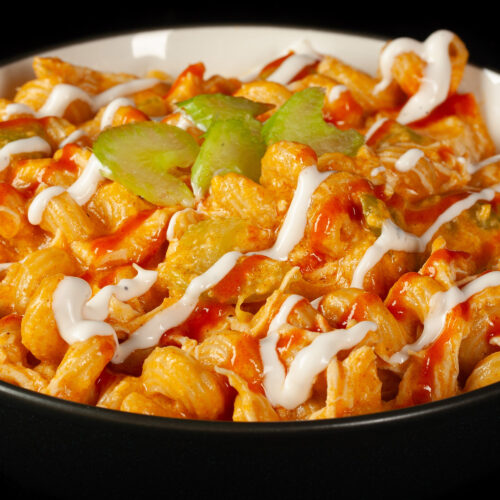 A bowl of buffalo chicken mac and cheese, with spiral pasta, cheese sauce, shredded chicken, celery, and drizzles of ranch dressing and frank's redhot sauce.
