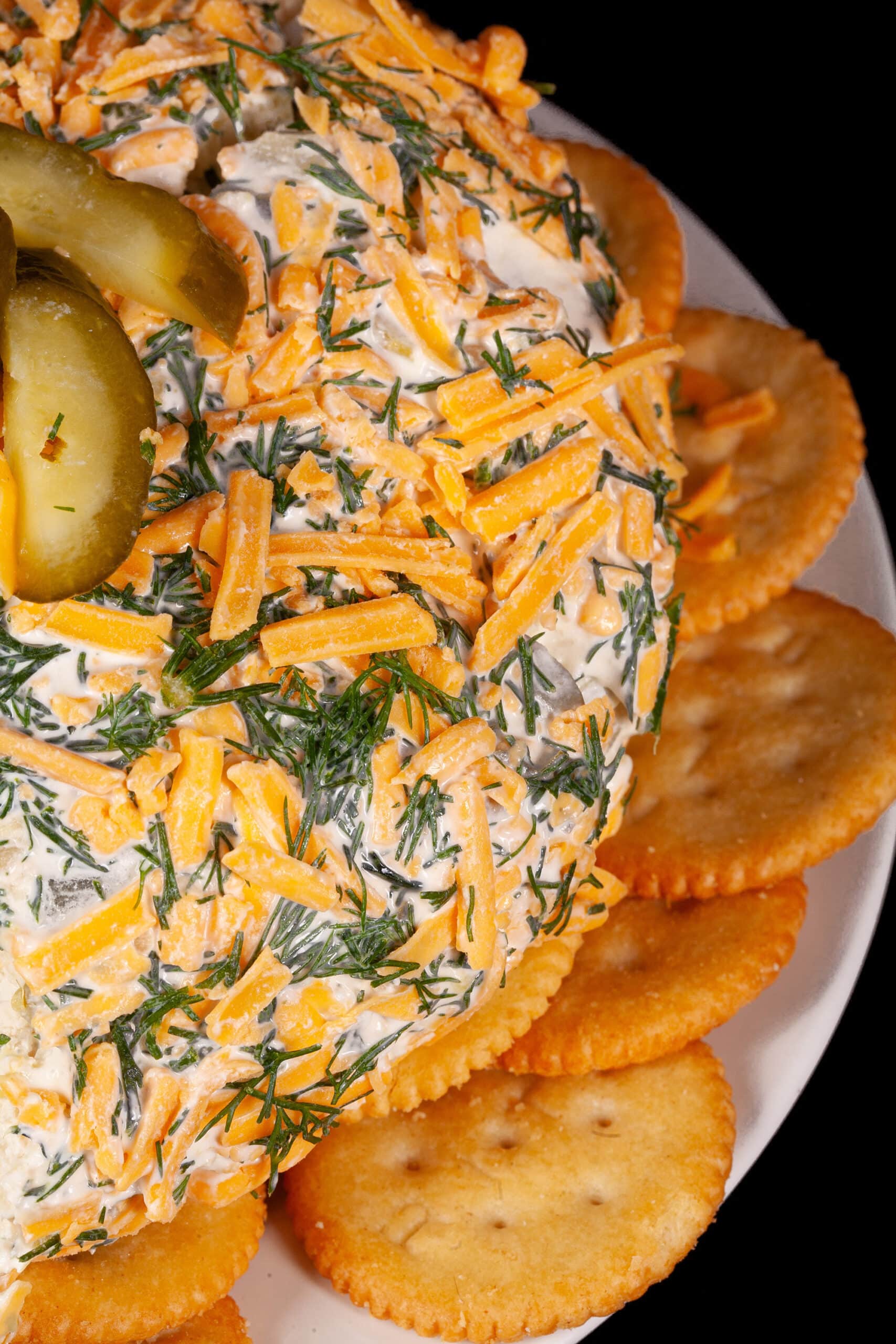 A round, cheese and dill covered dill pickle cheese ball on a plate with Ritz crackers.
