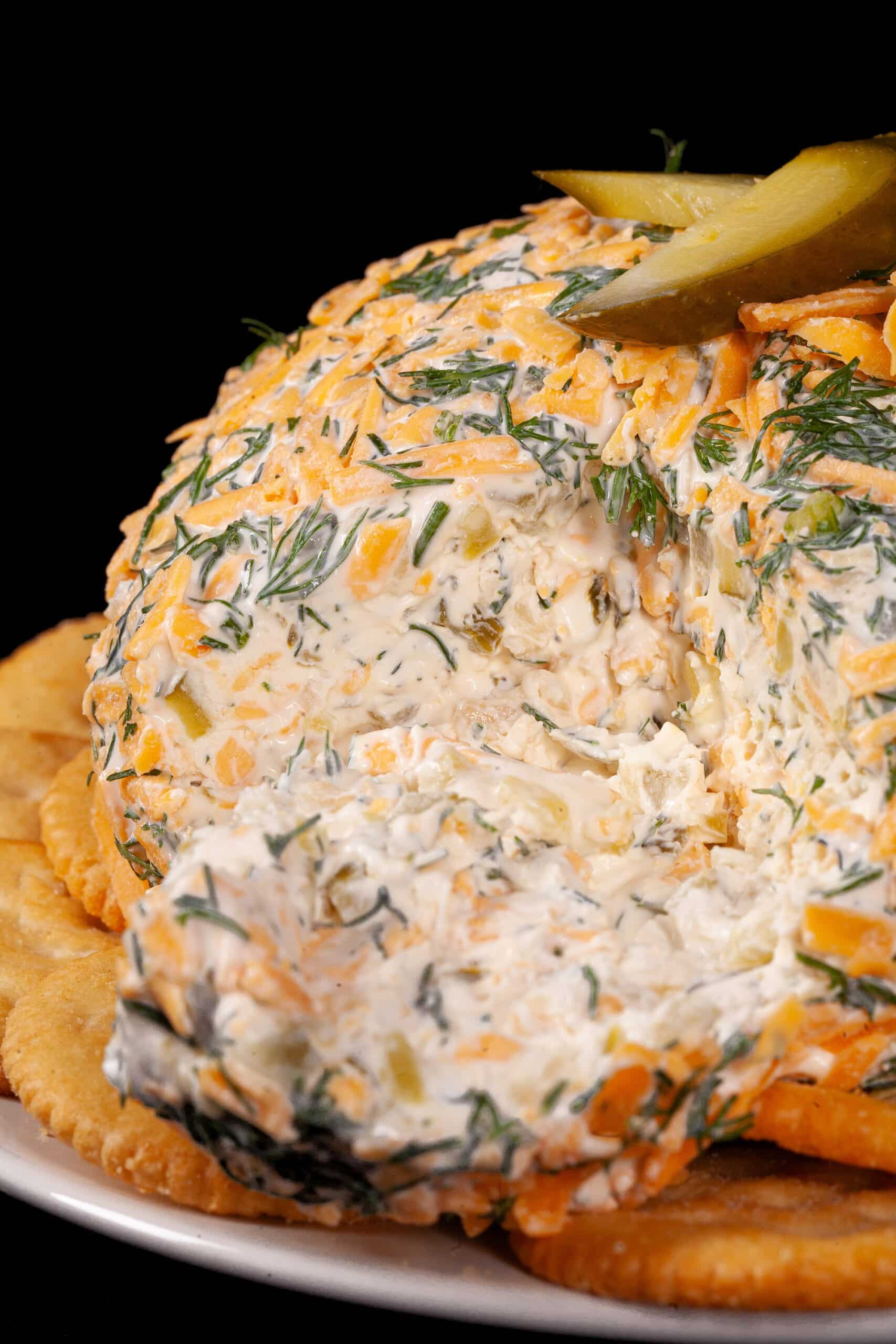 A round, cheese and dill covered dill pickle cheese ball on a plate with Ritz crackers.