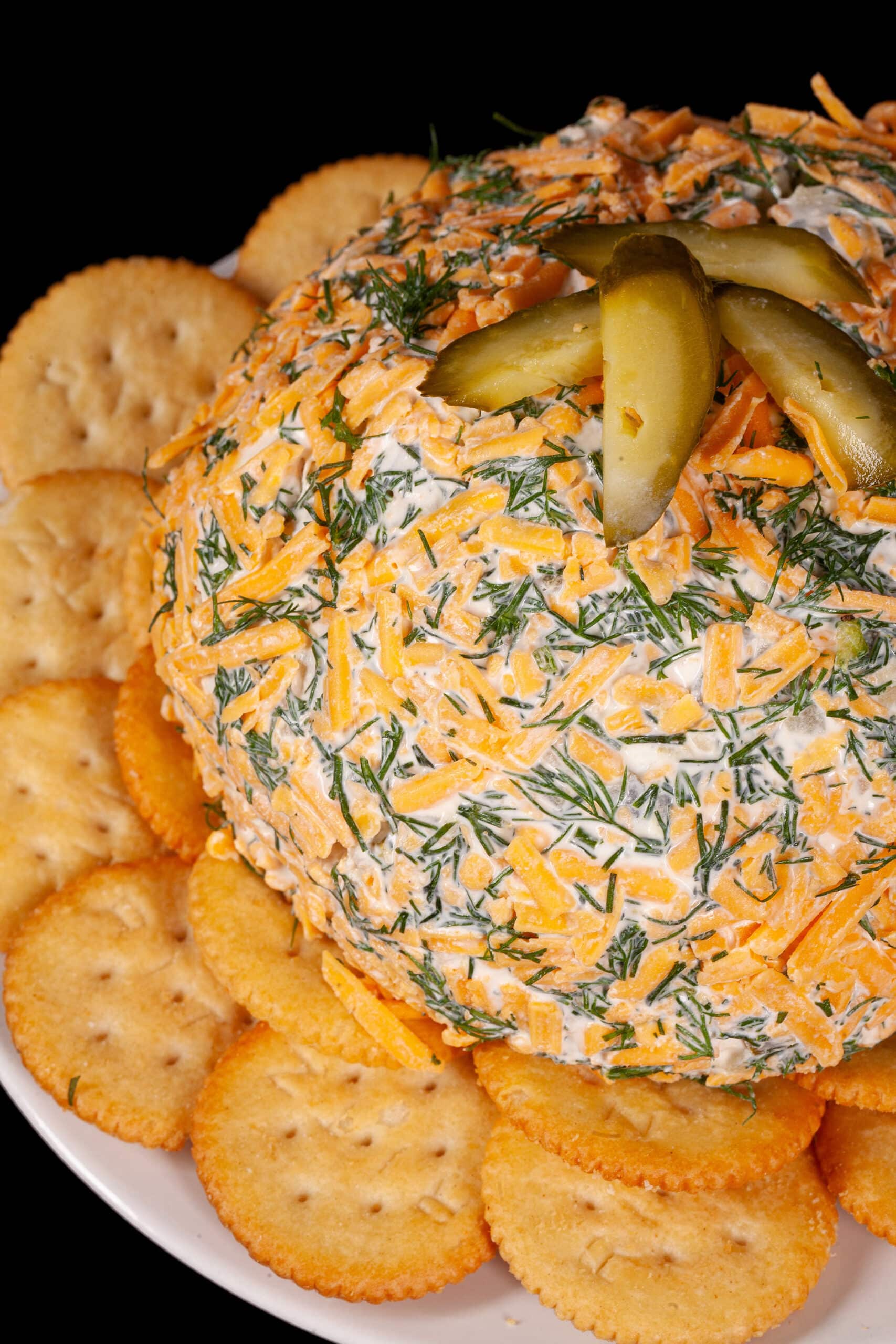 A round, cheese and dill covered dill pickle cheese ball on a plate with Ritz crackers.