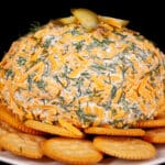 A round, cheese and dill covered dill pickle cheese ball on a plate with Ritz crackers.