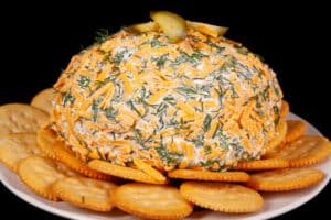 A round, cheese and dill covered dill pickle cheese ball on a plate with Ritz crackers.