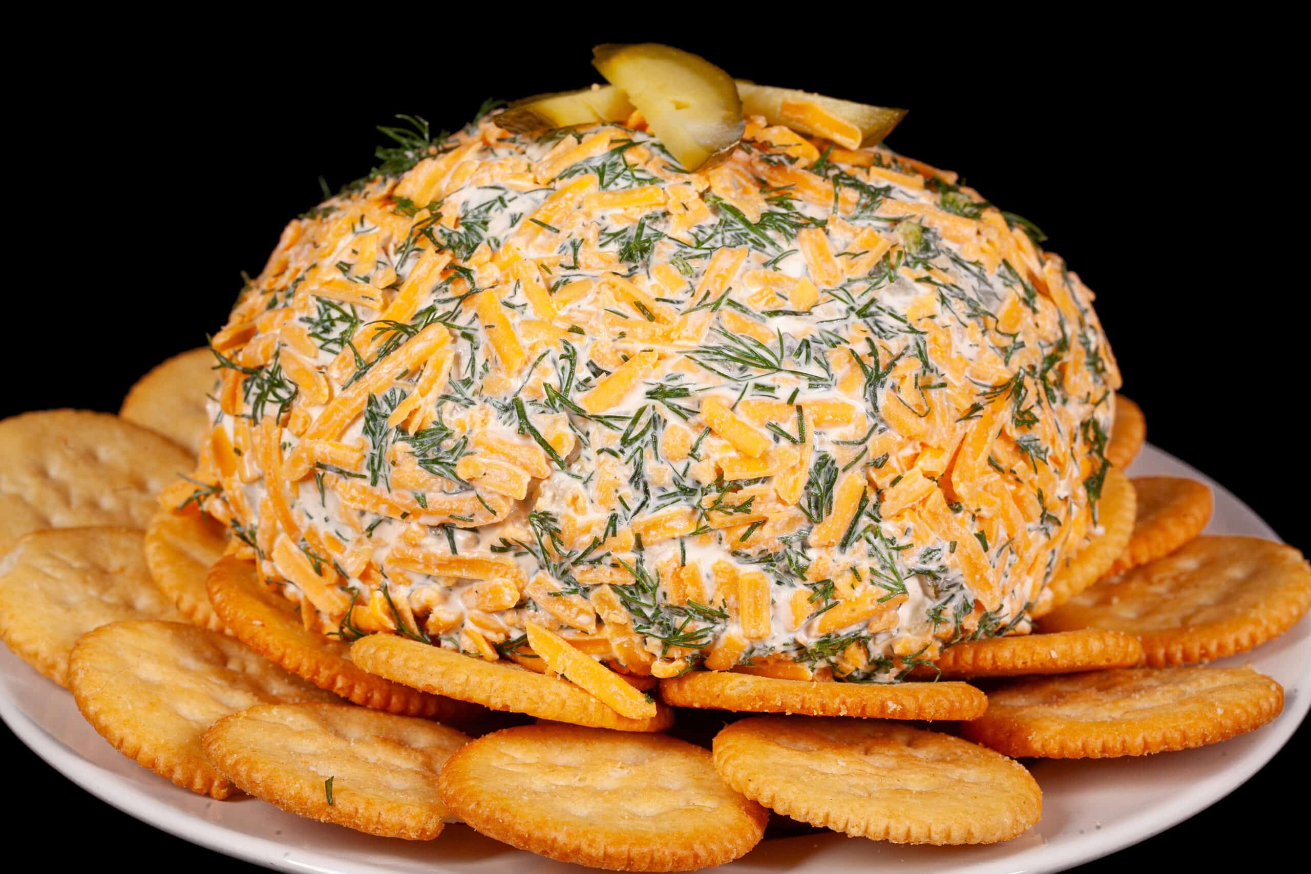 A round, cheese and dill covered dill pickle cheese ball on a plate with Ritz crackers.