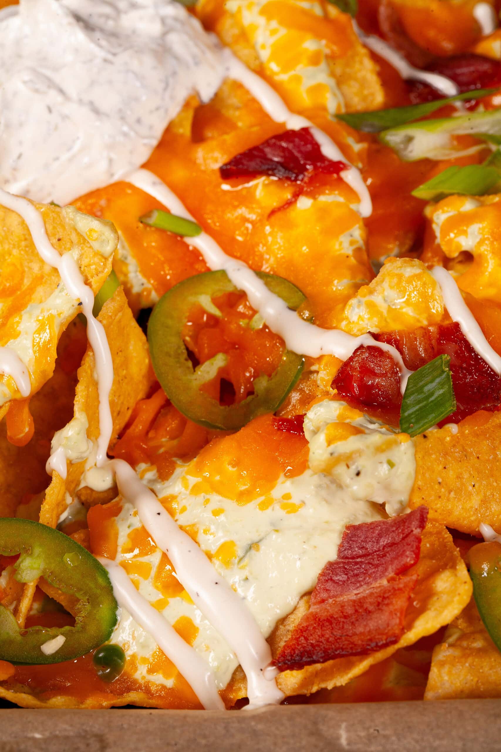 A pan of jalapeno popper nachos with jalapeno cream cheese queso, jalapeno slices, bacon, cheddar, green onions, ranch dressing, and ranch flavoured sour cream.
