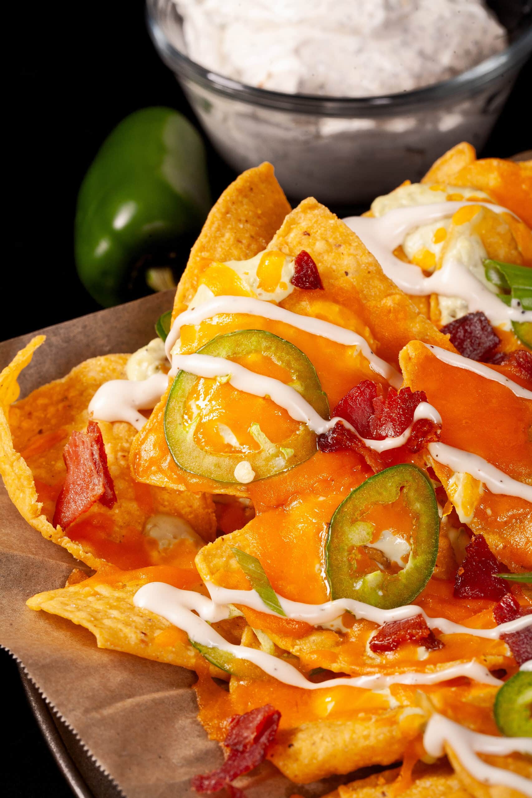 A pan of jalapeno popper nachos with jalapeno cream cheese queso, jalapeno slices, bacon, cheddar, green onions, ranch dressing, and ranch flavoured sour cream.