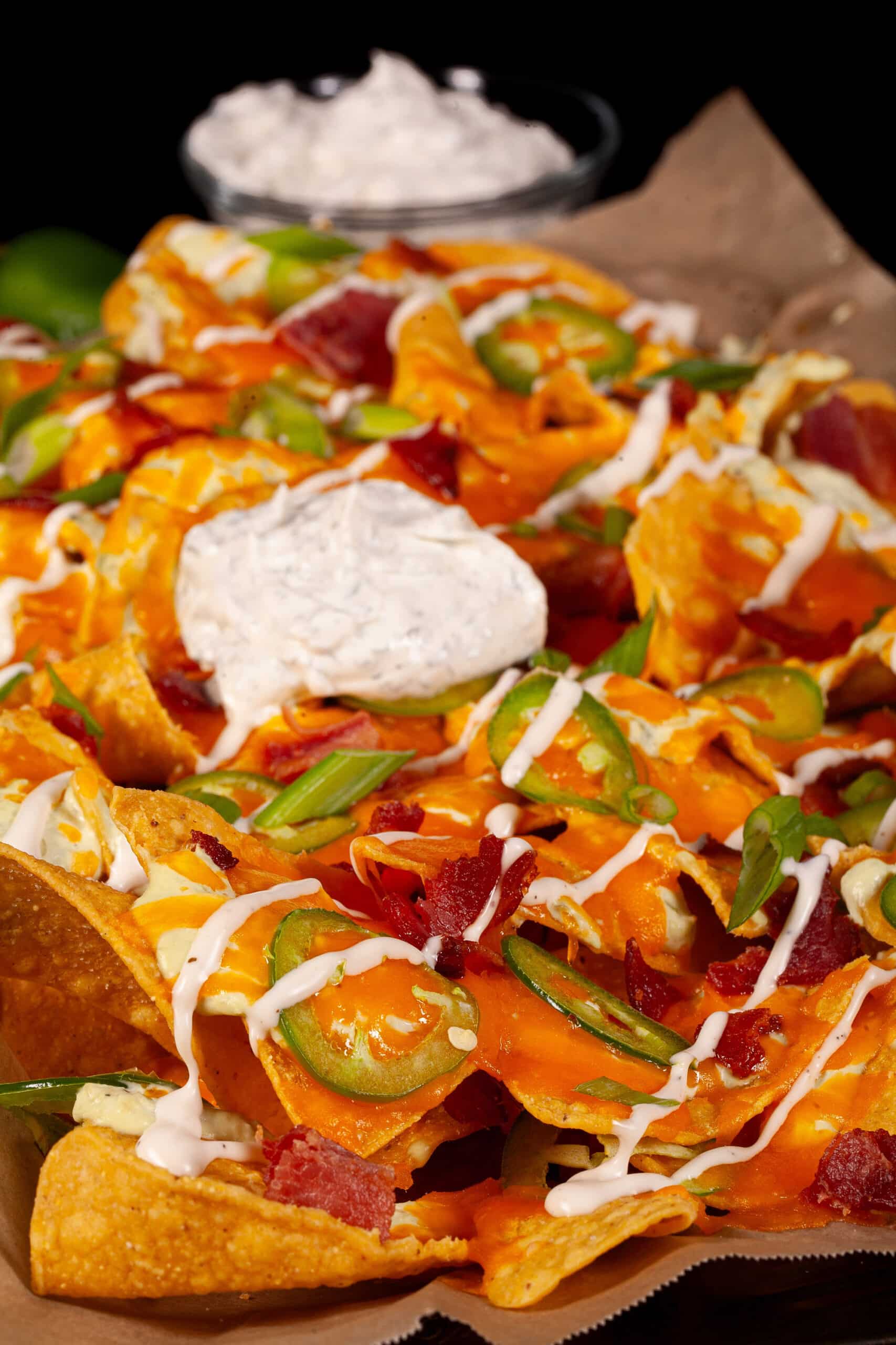 A pan of jalapeno popper nachos with jalapeno cream cheese queso, jalapeno slices, bacon, cheddar, green onions, ranch dressing, and ranch flavoured sour cream.