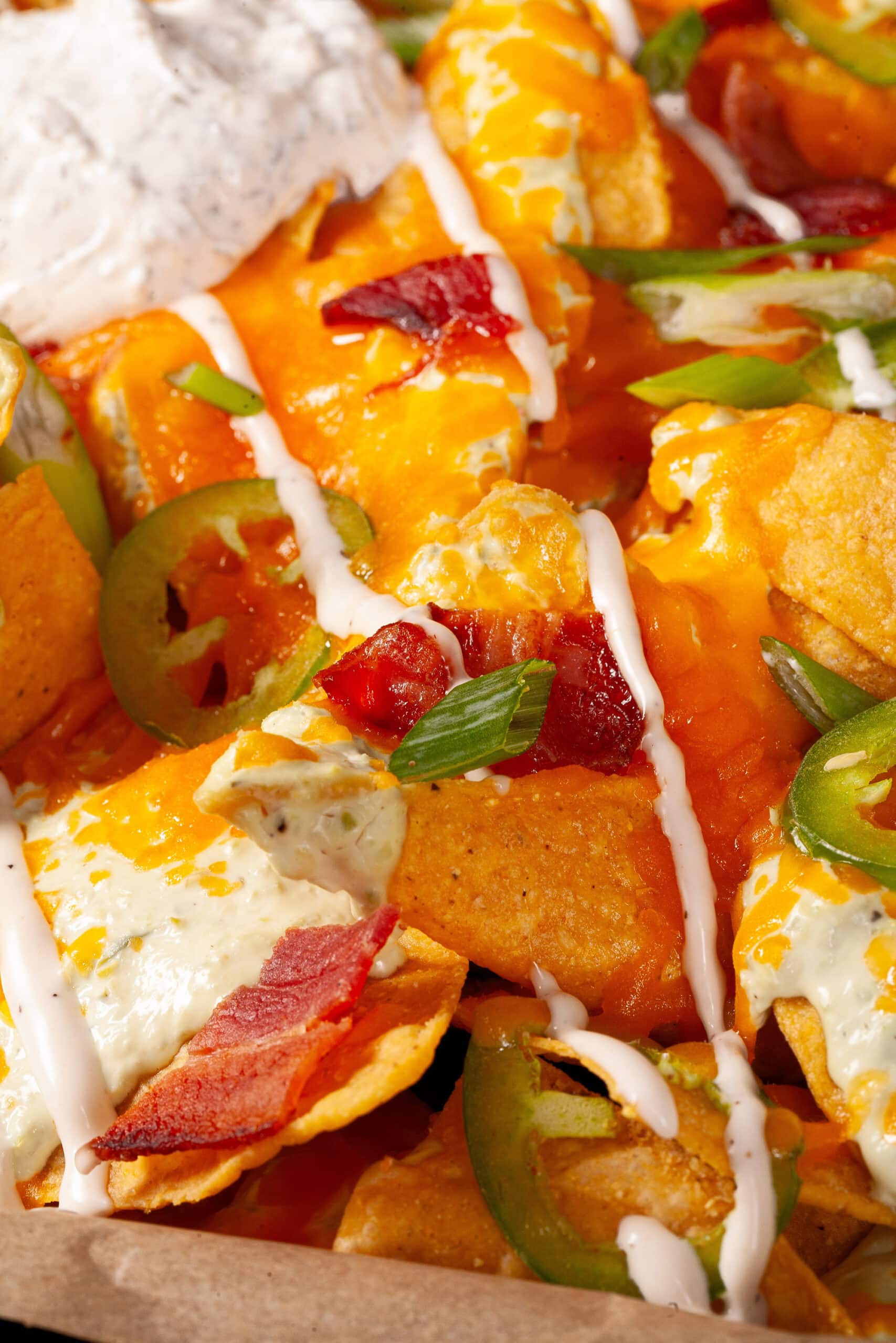 A pan of jalapeno popper nachos with jalapeno cream cheese queso, jalapeno slices, bacon, cheddar, green onions, ranch dressing, and ranch flavoured sour cream.