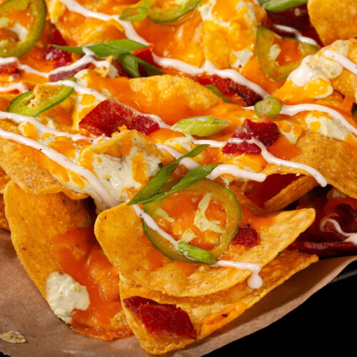 A pan of jalapeno popper nachos with jalapeno cream cheese queso, jalapeno slices, bacon, cheddar, green onions, ranch dressing, and ranch flavoured sour cream.