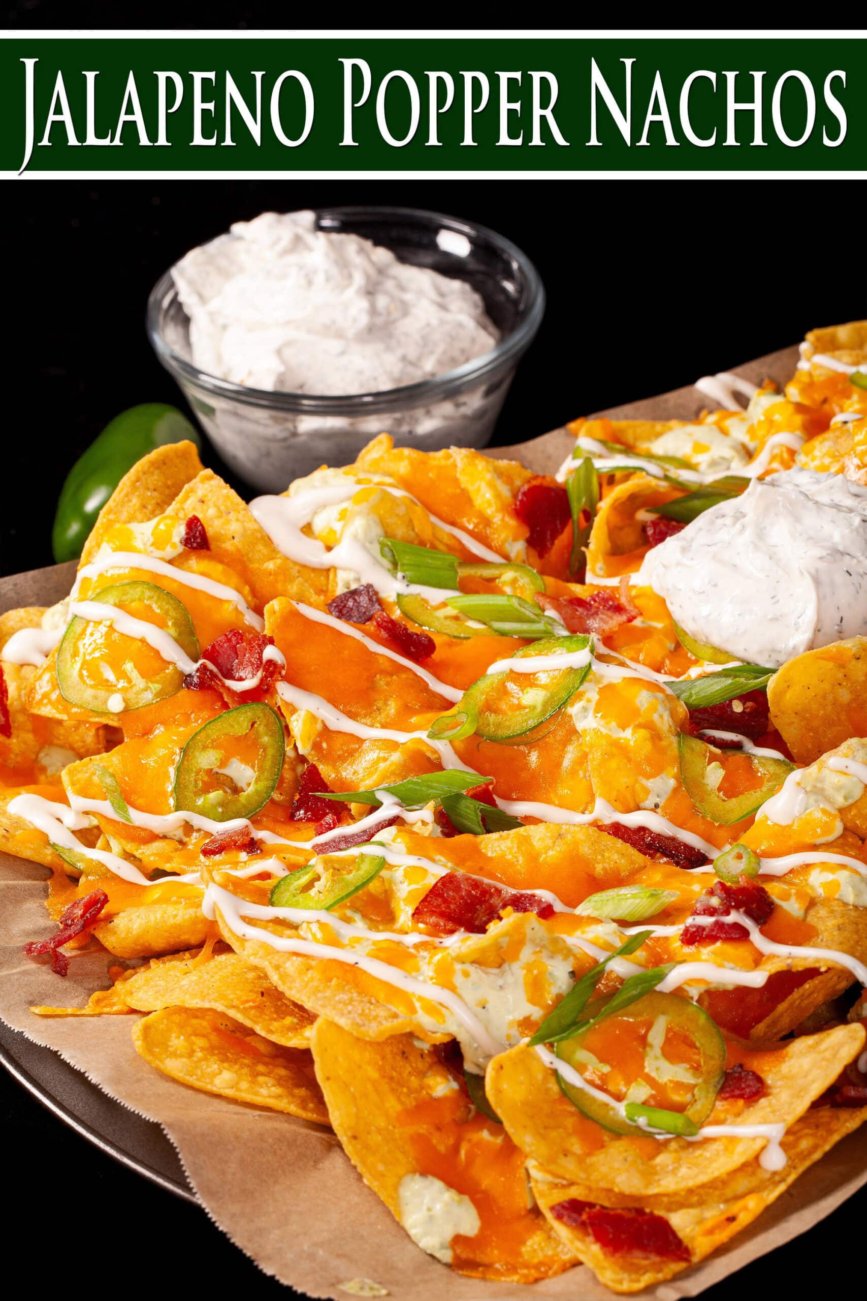 A pan of jalapeno popper nachos with jalapeno cream cheese queso, jalapeno slices, bacon, cheddar, green onions, ranch dressing, and ranch flavoured sour cream. Overlaid text says jalapeno popper nachos.