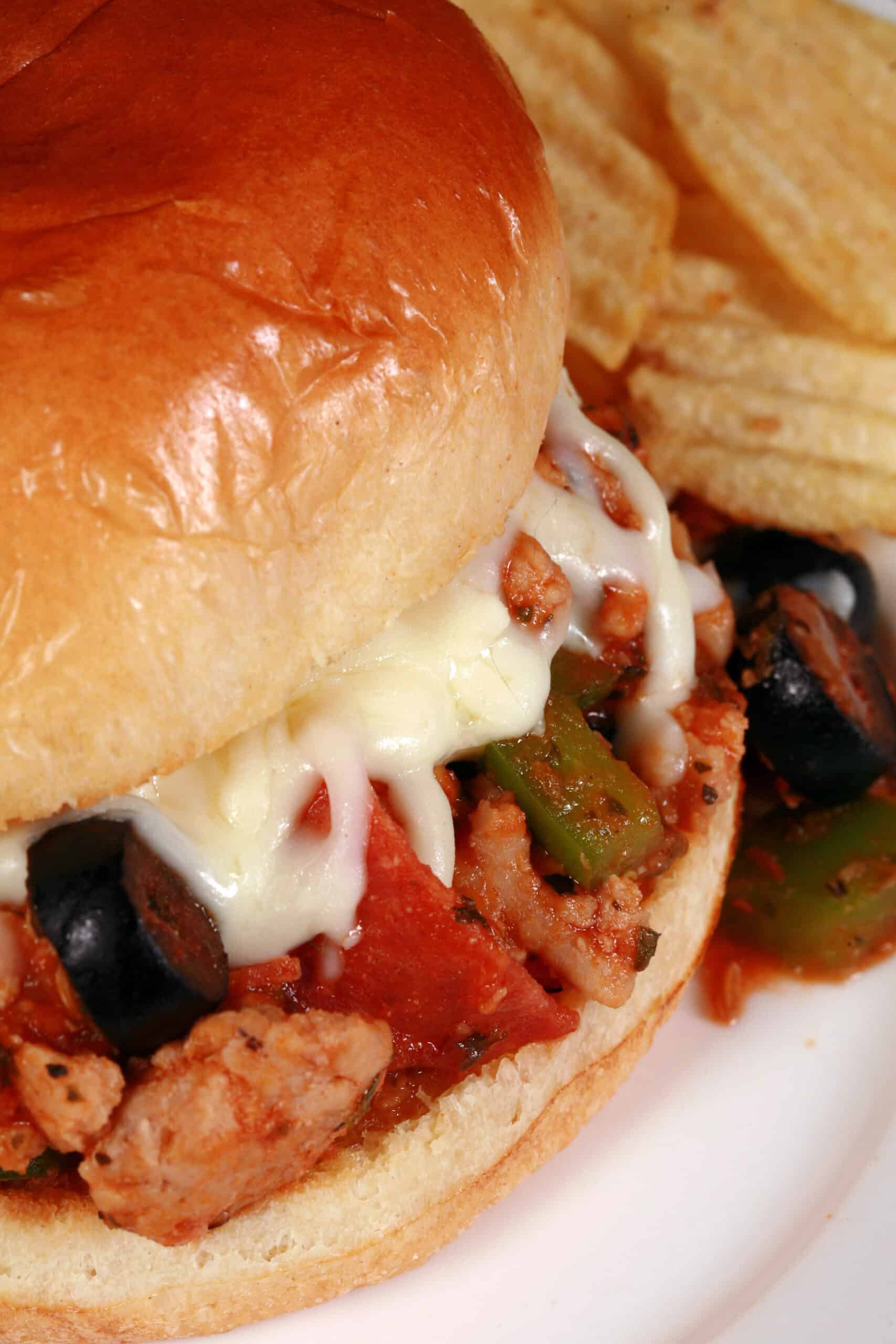 A pizza themed sloppy joes sandwich on a plate with chips, with pepperoni, green pepper, black olives, and mozzarella cheese visible.