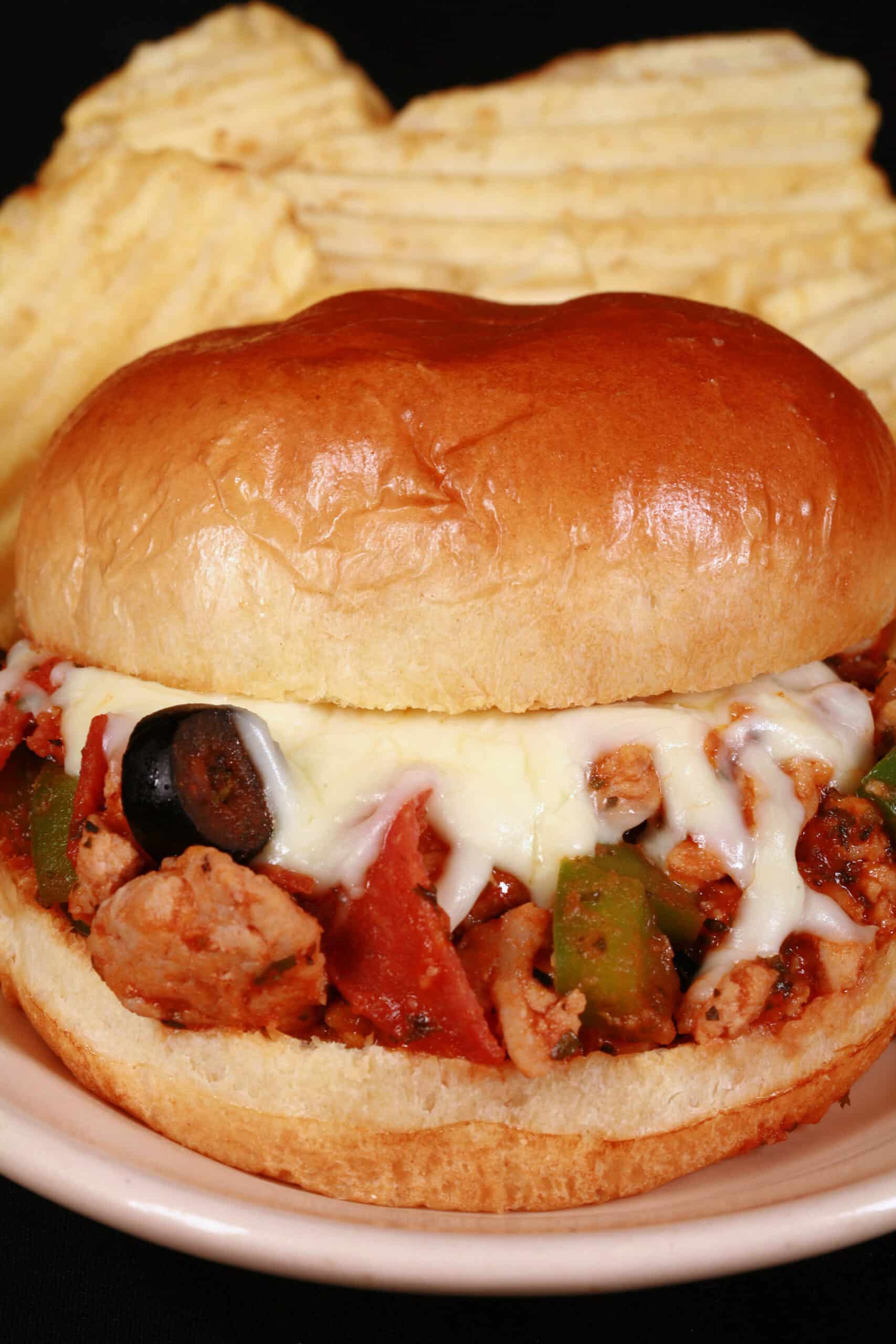 A pizza sloppy joes sandwich on a plate with chips, with pepperoni, green pepper, black olives, and mozzarella cheese visible.