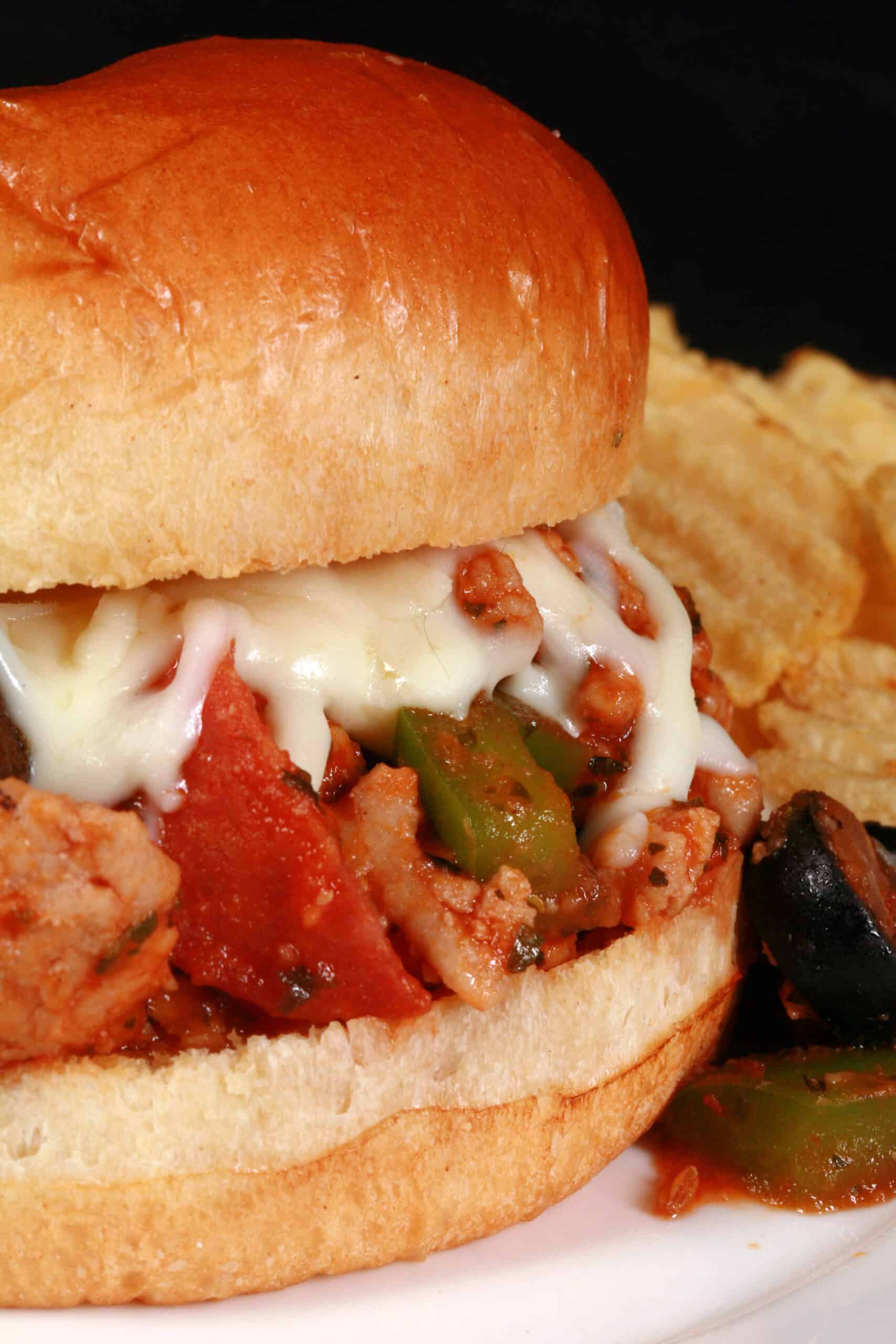 A pizza themed sloppy joes sandwich on a plate with chips, with pepperoni, green pepper, black olives, and mozzarella cheese visible.
