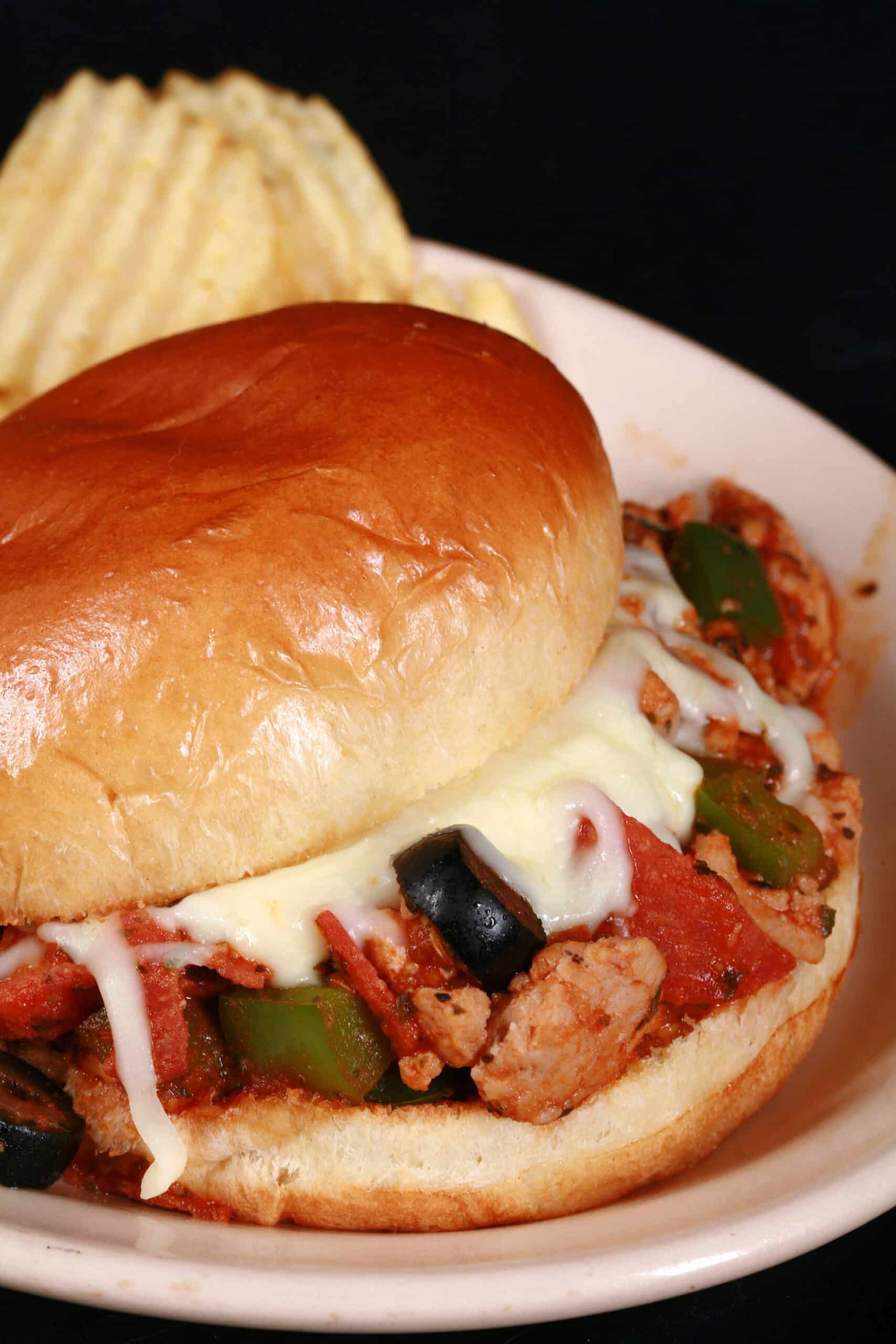 A pizza sloppy joes sandwich on a plate with chips, with pepperoni, green pepper, black olives, and mozzarella cheese visible.