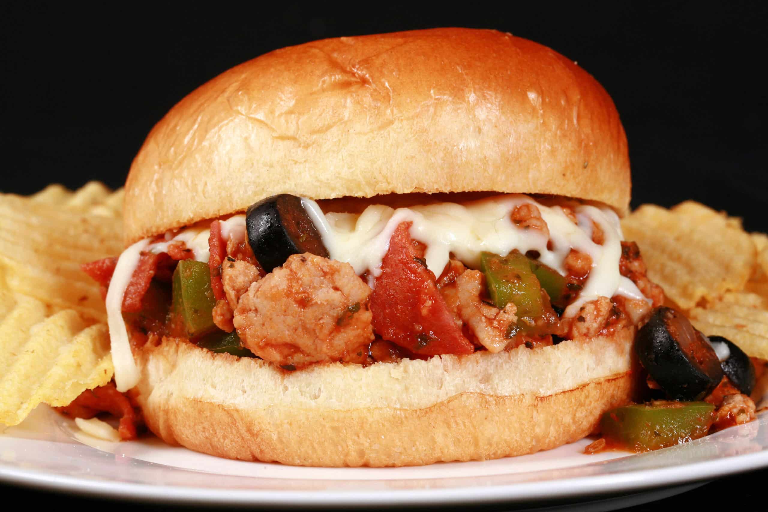 A pizza themed sloppy joes sandwich on a plate with chips, with pepperoni, green pepper, black olives, and mozzarella cheese visible.