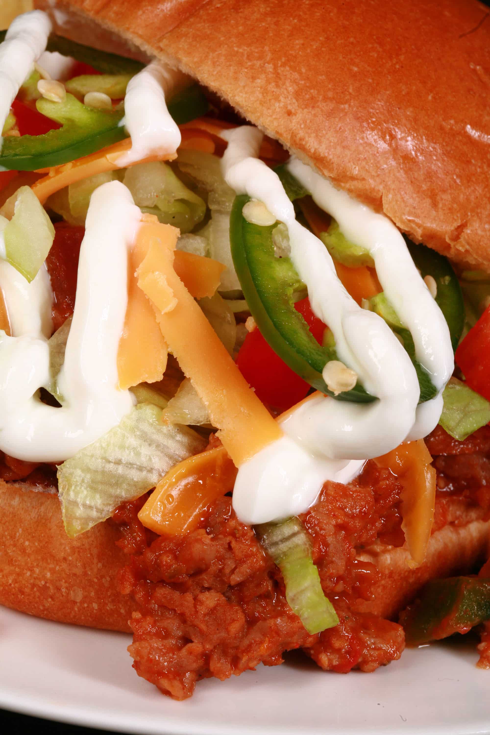 A Taco sloppy joes sandwich, with taco meat, lettuce, cheese, salsa, sour cream, and jalapeno slices, on a plate with tortilla chips.