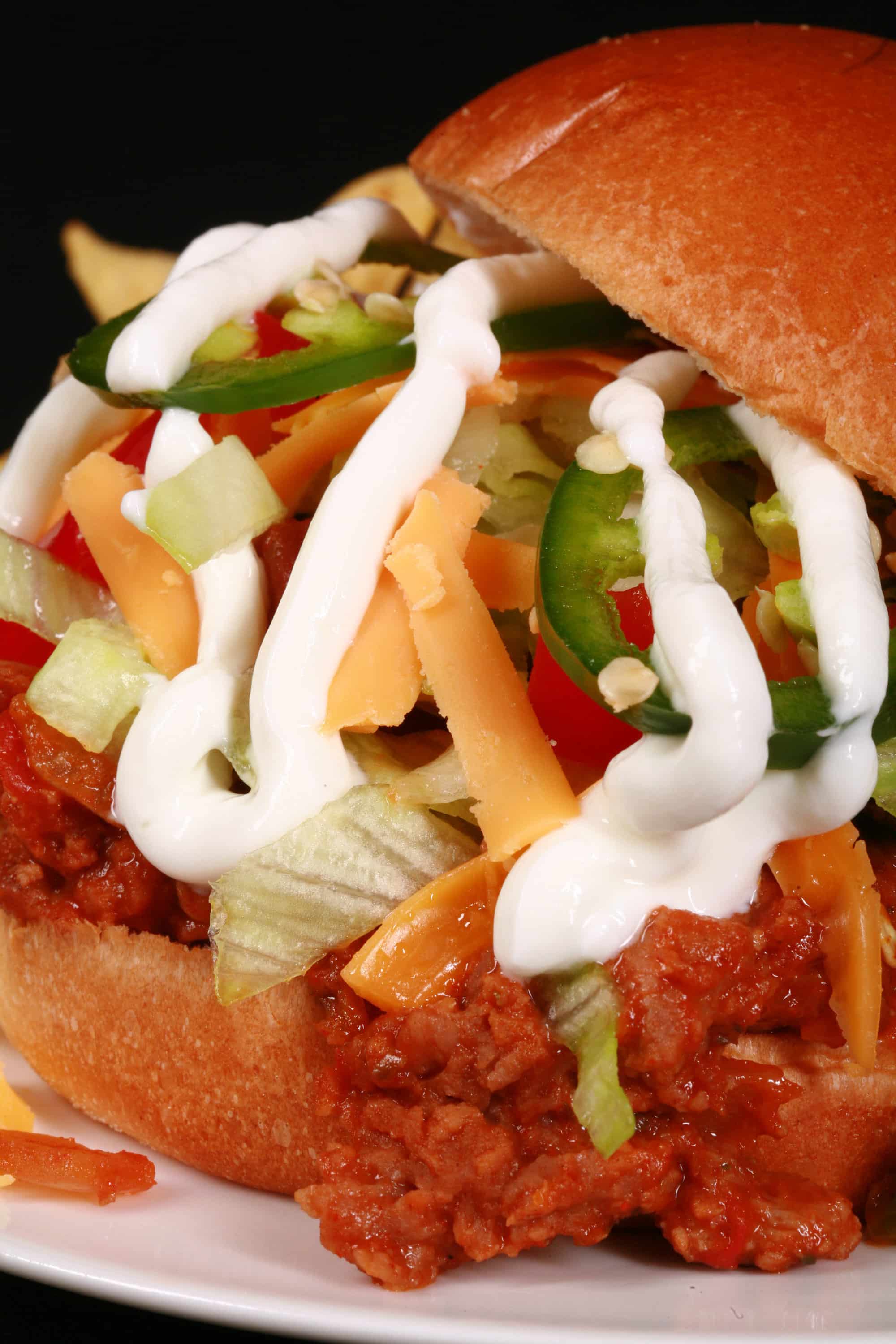 A Taco themed sloppy joe sandwich, with taco meat, lettuce, cheese, salsa, sour cream, and jalapeno slices, on a plate with tortilla chips.